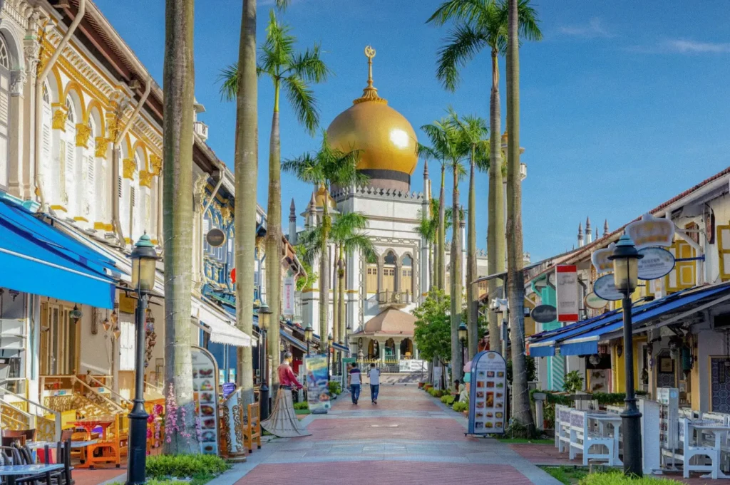Wide-angle street-level shot of Kampong Glam in Singapore, with palm-lined pedestrian walkway, colorful heritage shophouses, outdoor restaurant seating, and the golden dome of Sultan Mosque in the background under a blue sky.