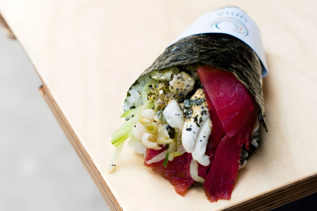 Close-up of a sushi hand roll on a wooden surface. Seaweed wrap holds white rice, vibrant pink tuna slices, and green vegetables, creating a fresh, inviting look.
