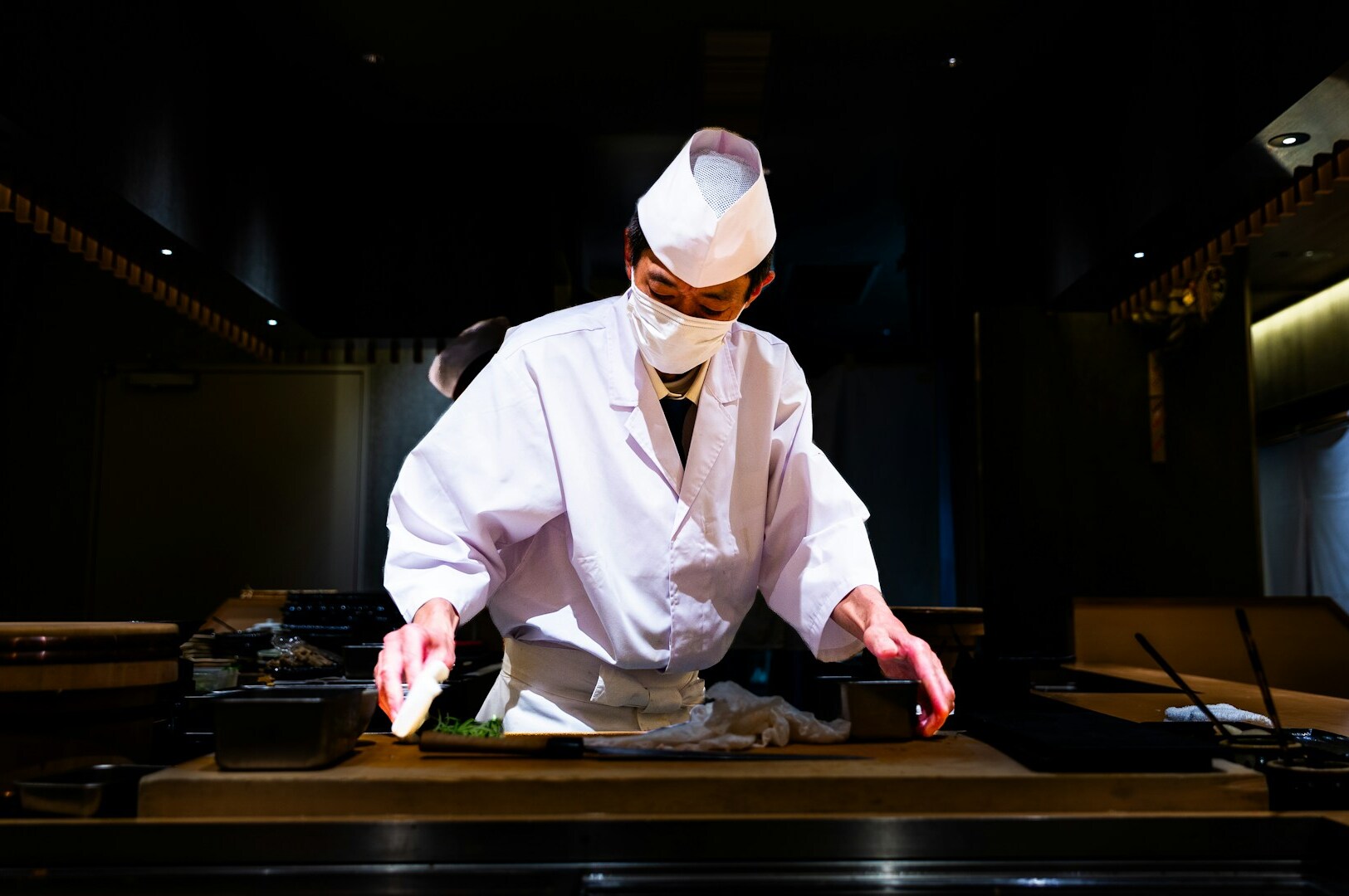 A chef in a white coat and mask skillfully prepares food in a highly anticipated celebrity chef restaurant setting.
