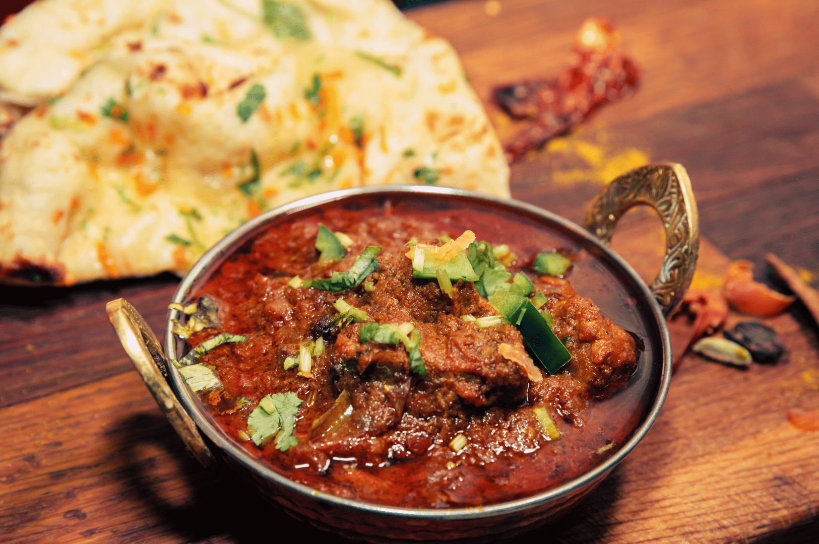 Rich red Indian curry with tender meat chunks, garnished with herbs and chilies, served alongside naan—evoking the bold, rustic flavors of Little India’s spice-forward dining experiences.
