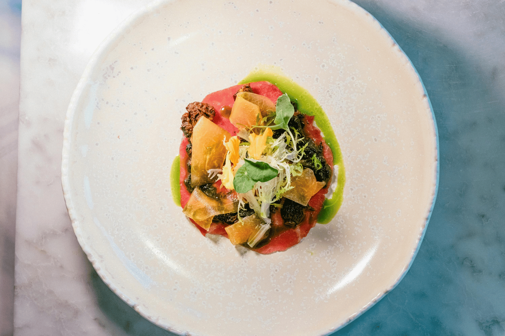 Elegant plant-based appetizer at a fine dining vegan restaurant in Singapore, featuring beetroot carpaccio with microgreens, crispy chips, and herb puree on a speckled ceramic plate.