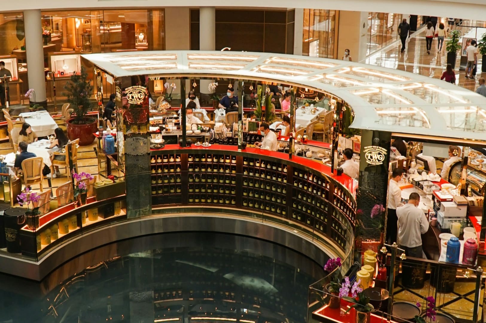 A bustling indoor restaurant with elegant gold and wood decor, seated patrons, and busy staff. It overlooks a reflective water feature, enhancing its upscale ambiance.