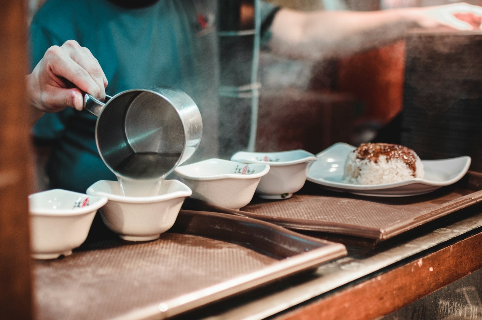 A person pours hot water into a bowl at a bustling Singapore hawker center, preparing a dish.
