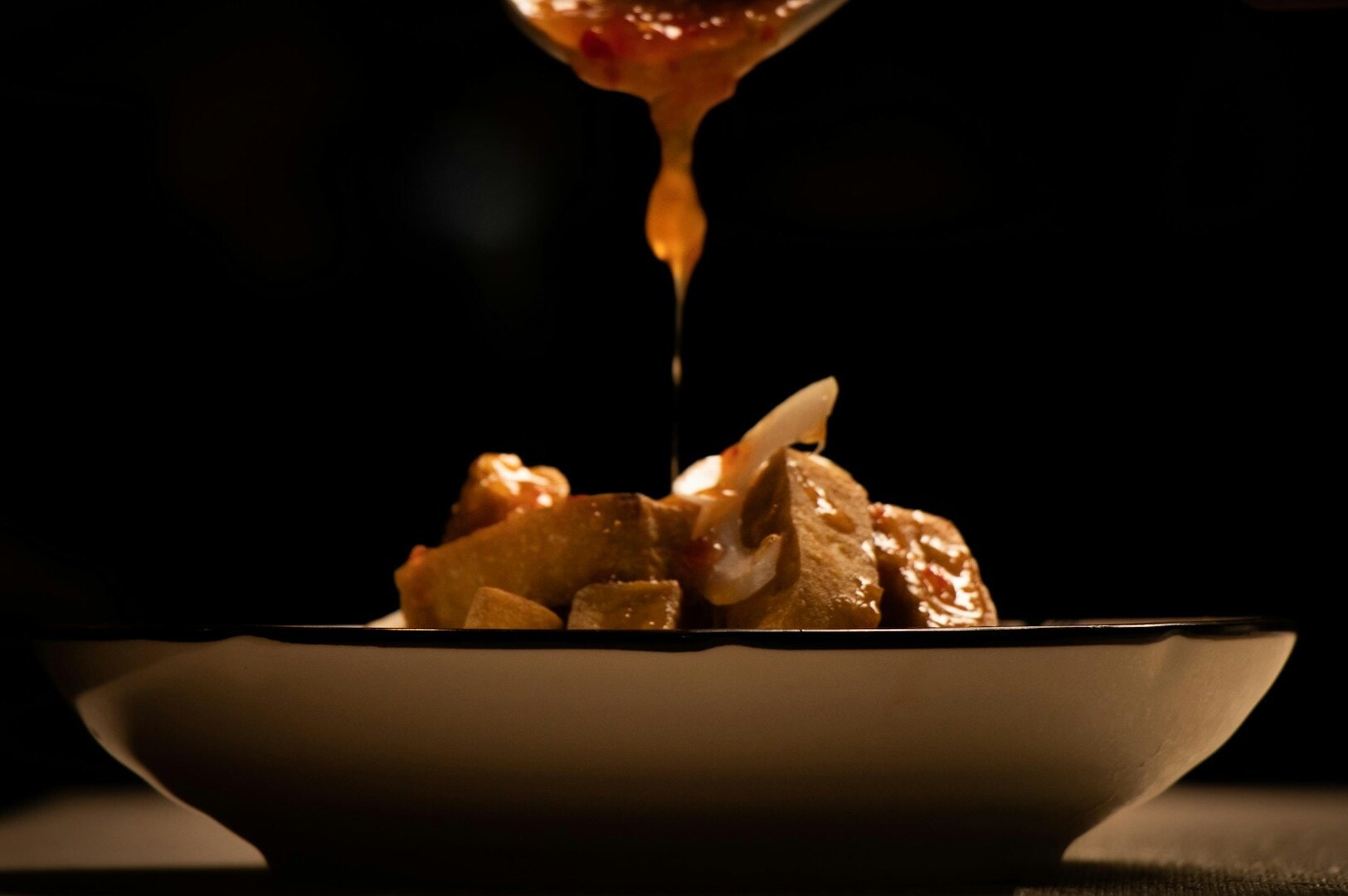 Rich sauce drizzles over a plate of tofu and vegetables, set on a dark background. The lighting highlights a warm, appetizing scene.