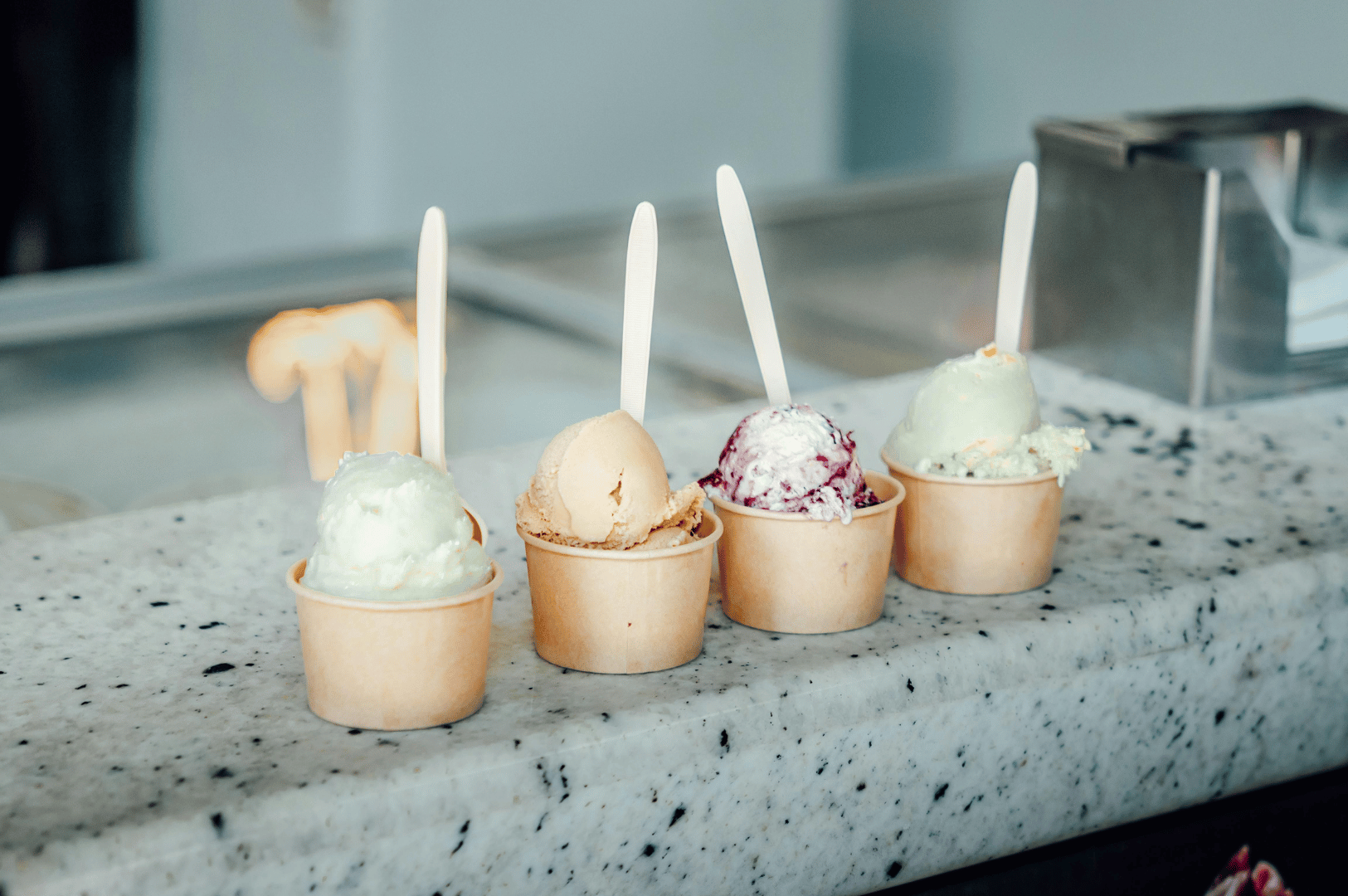 Assorted vegan ice cream scoops at a Singapore dessert shop, featuring unique plant-based flavors in paper cups with wooden spoons on a granite countertop.