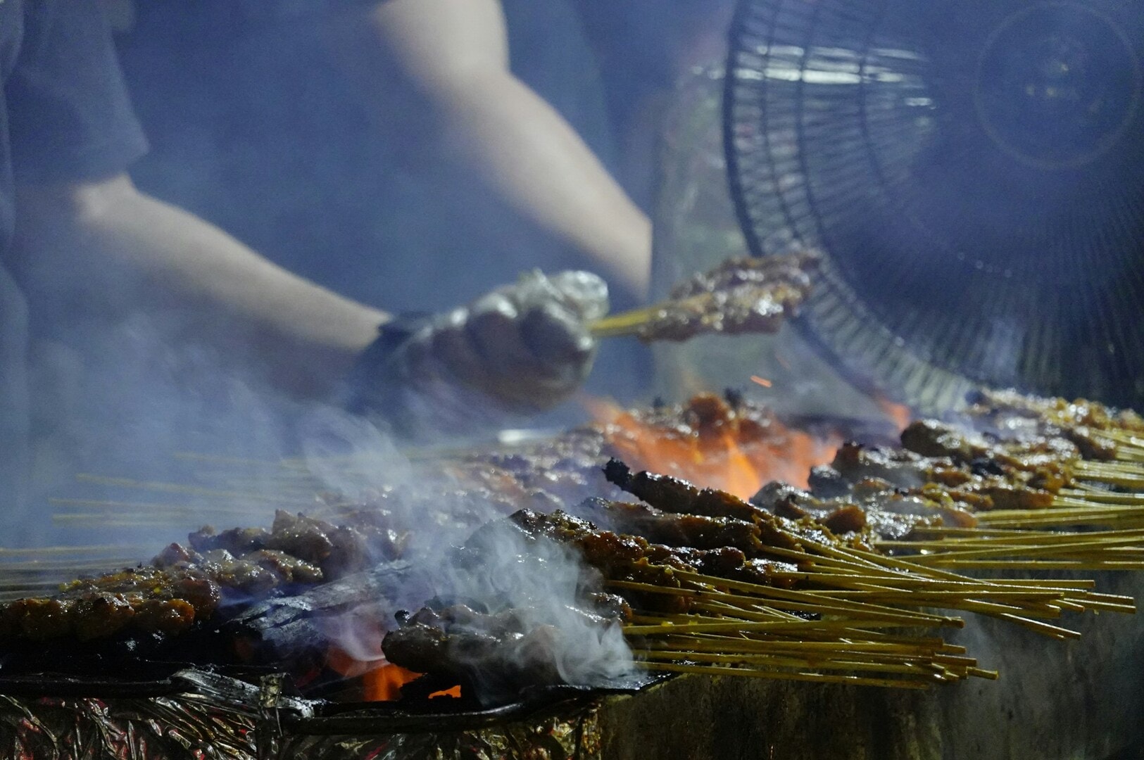A man cooks skewered meat over a grill while a fan circulates air in a bustling Singapore hawker center.