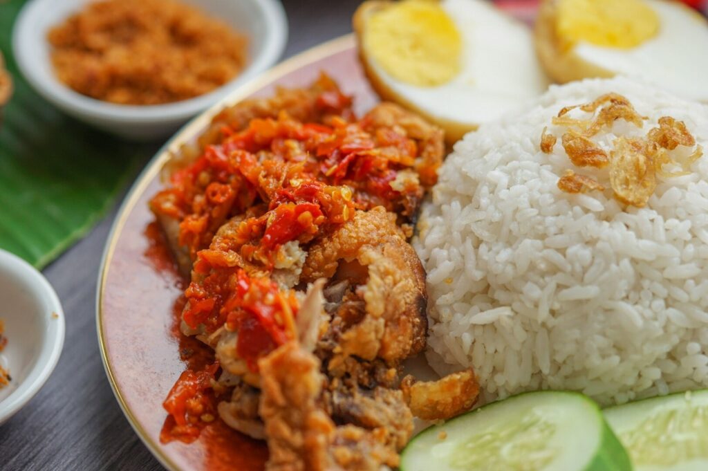 A plate featuring spicy fried chicken topped with red chili sauce, white rice garnished with crispy shallots, cucumber slices, and halved boiled egg.