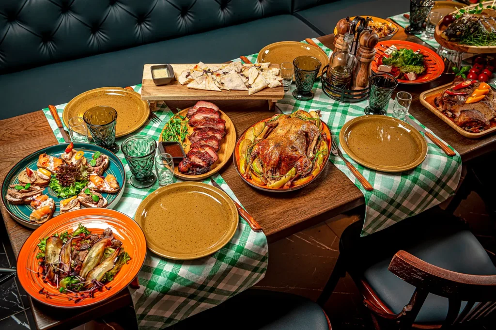 Table filled with assorted dishes including roasted meats, salads, grilled seafood, and side plates, arranged on a checkered tablecloth for a restaurant dining spread.