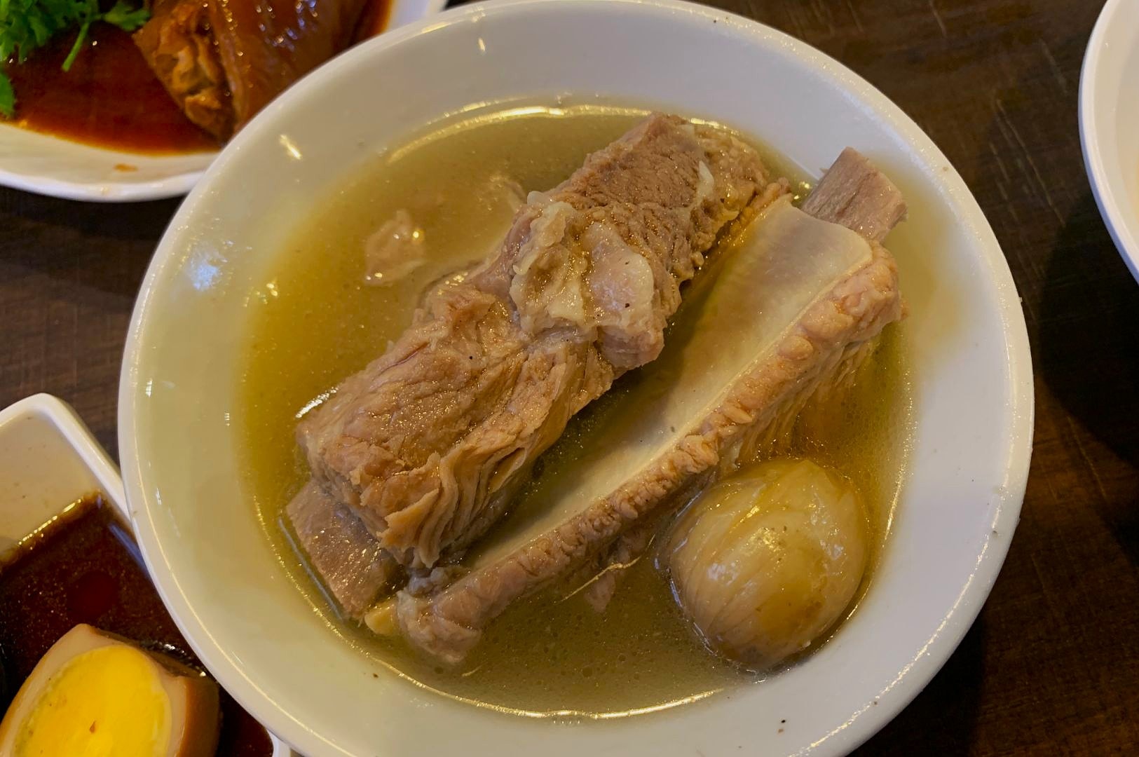 A bowl of light-colored broth with tender pork ribs and a whole onion submerged. Nearby are partially visible plates of sauce and braised meat.