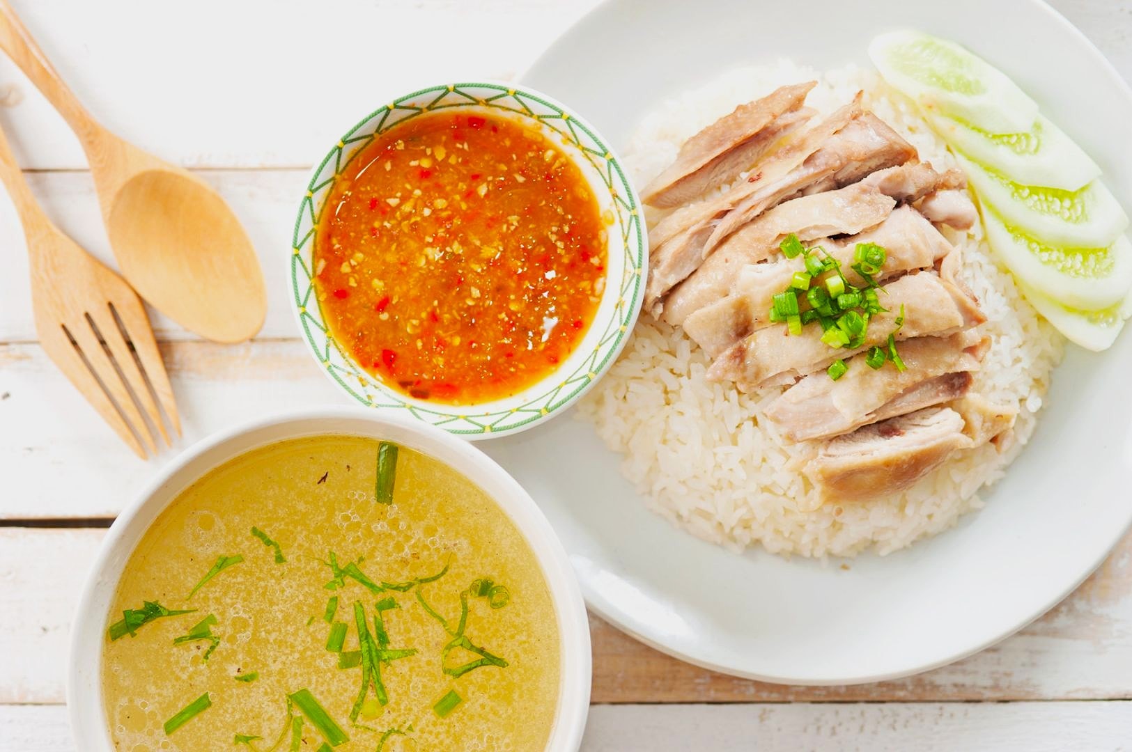 Plate of sliced chicken on rice garnished with green onions and cucumber slices, with a side of clear soup with herbs and a spicy dipping sauce. Wooden cutlery rests nearby.