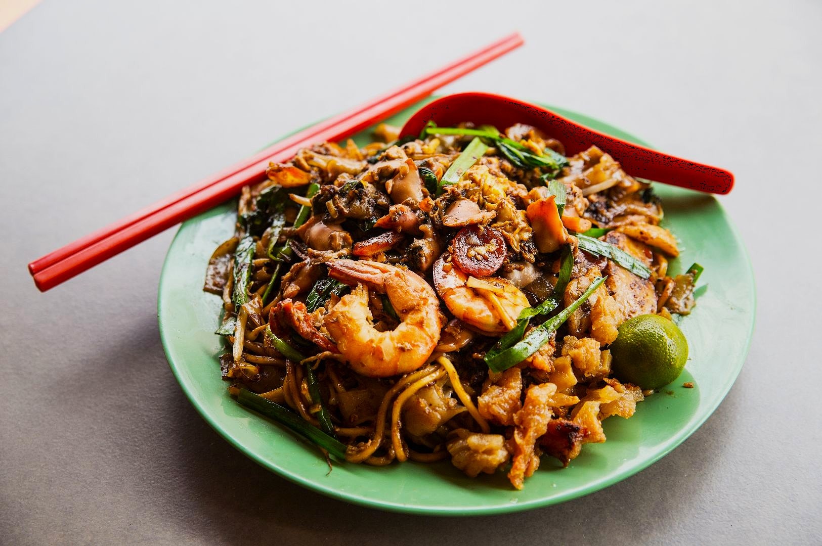 A vibrant plate of char kway teow with shrimp, noodles, and vegetables on a green plate, accompanied by red chopsticks and a lime, exuding a savory aroma.