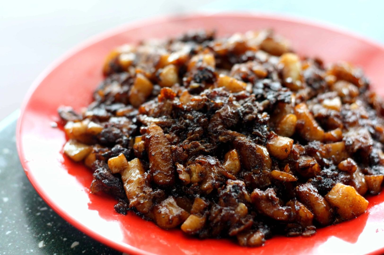 A vibrant red plate filled with dark, caramelized chunks of fried radish cake glistening with oil, showcasing a savory and rich appearance.