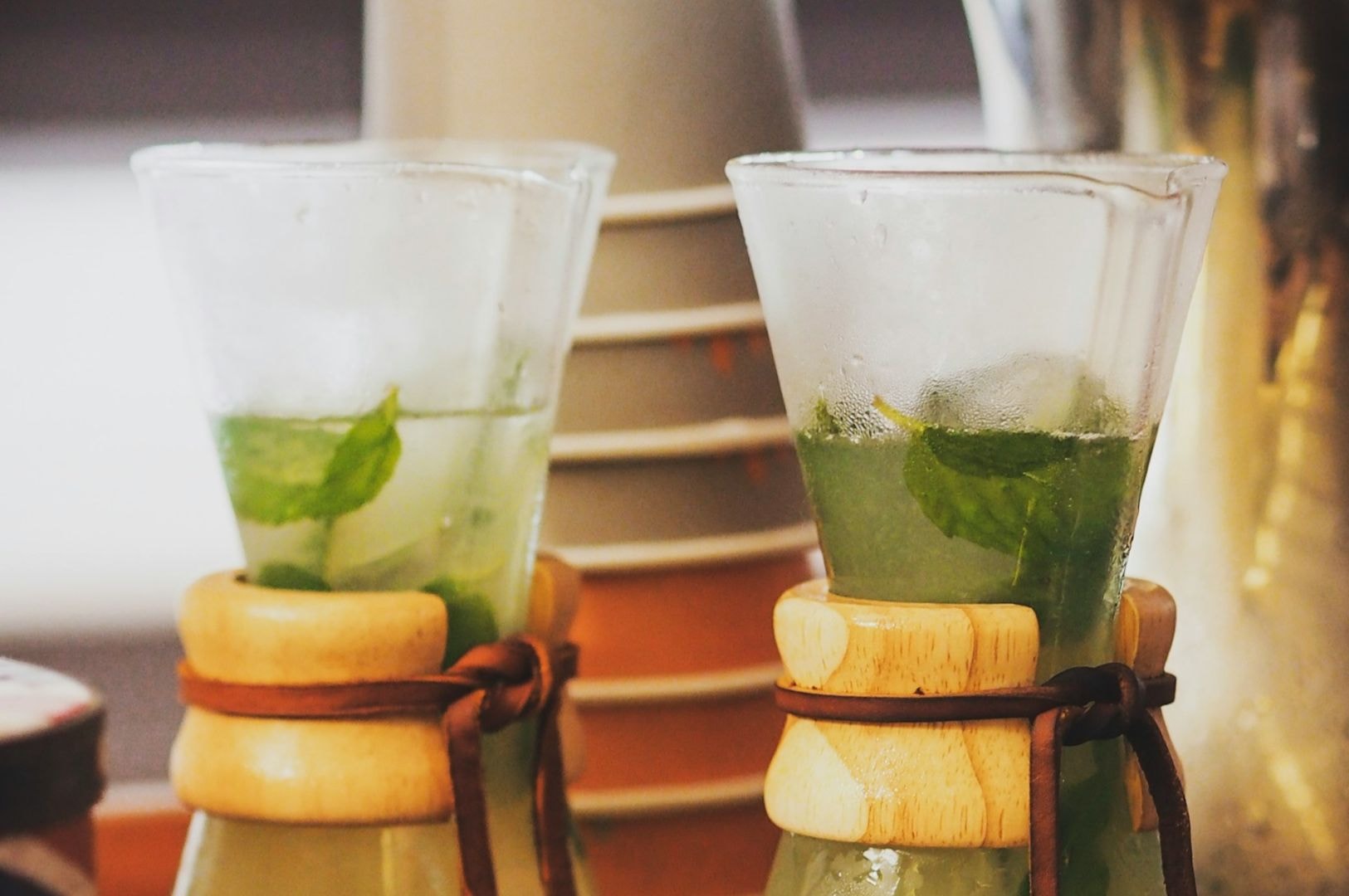 Close-up of two frosty glasses filled with mint leaves and a light green beverage. Glasses have wooden necks with leather ties. Background includes stacked cups.