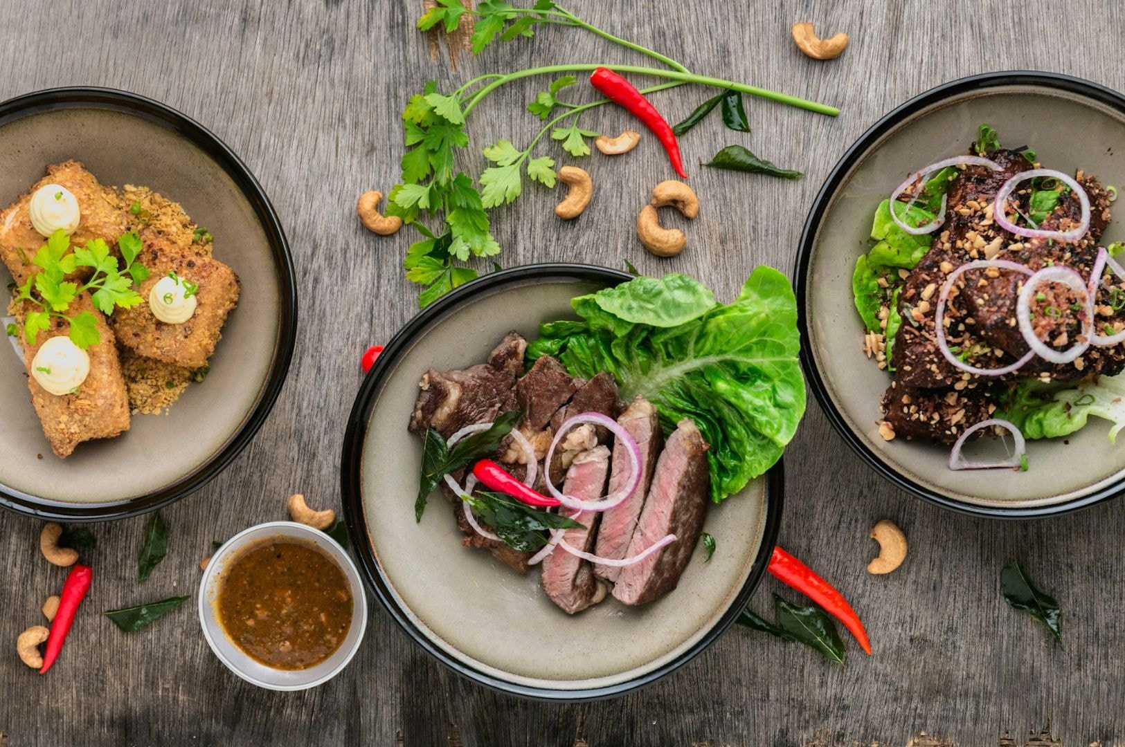 Three dishes on a wooden table; crispy rolls with greens, sliced steak with lettuce, and a seed-crusted salad. Cashews and chilies garnish the scene.