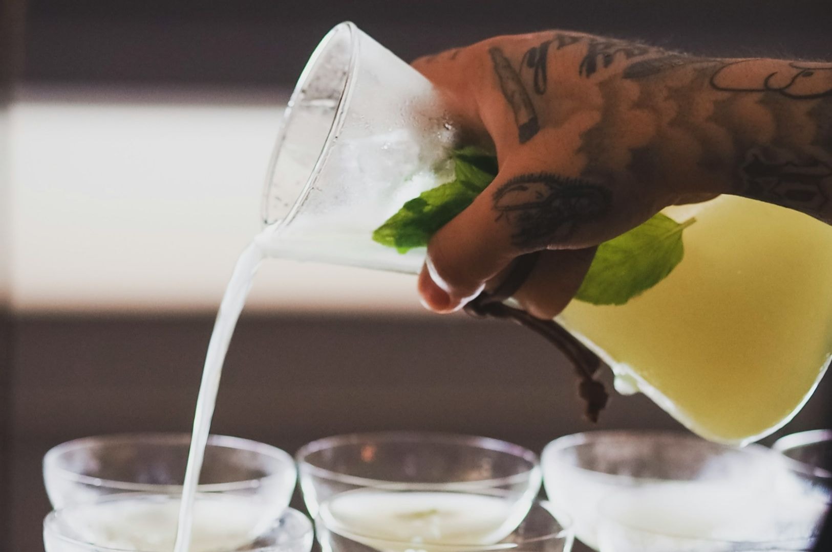 A tattooed hand pours a light green liquid with mint leaves from a glass pitcher into several elegant cocktail glasses, evoking a refreshing and artistic ambiance.