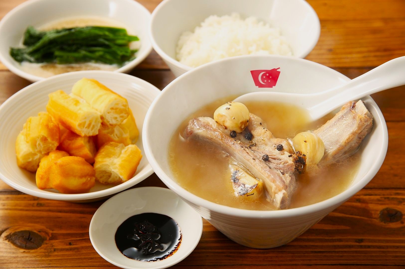 A steaming bowl of bak kut teh with ribs and garlic is in the foreground, accompanied by bowls of rice, youtiao, spinach, and soy sauce. Warm, inviting tone.