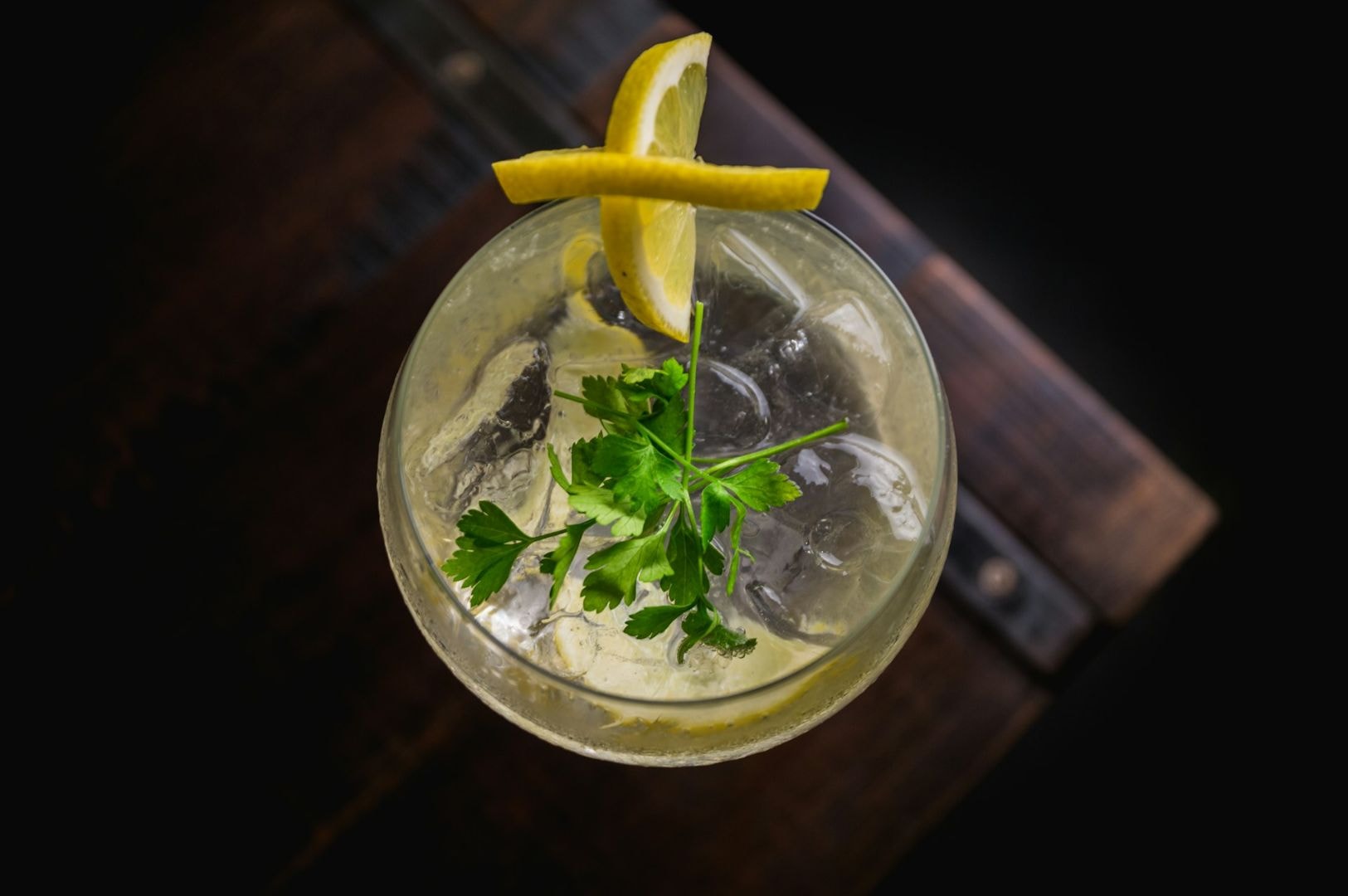 Top-down view of a cocktail in a glass with ice, garnished with fresh parsley and a lemon twist, set on a rustic dark wood surface.