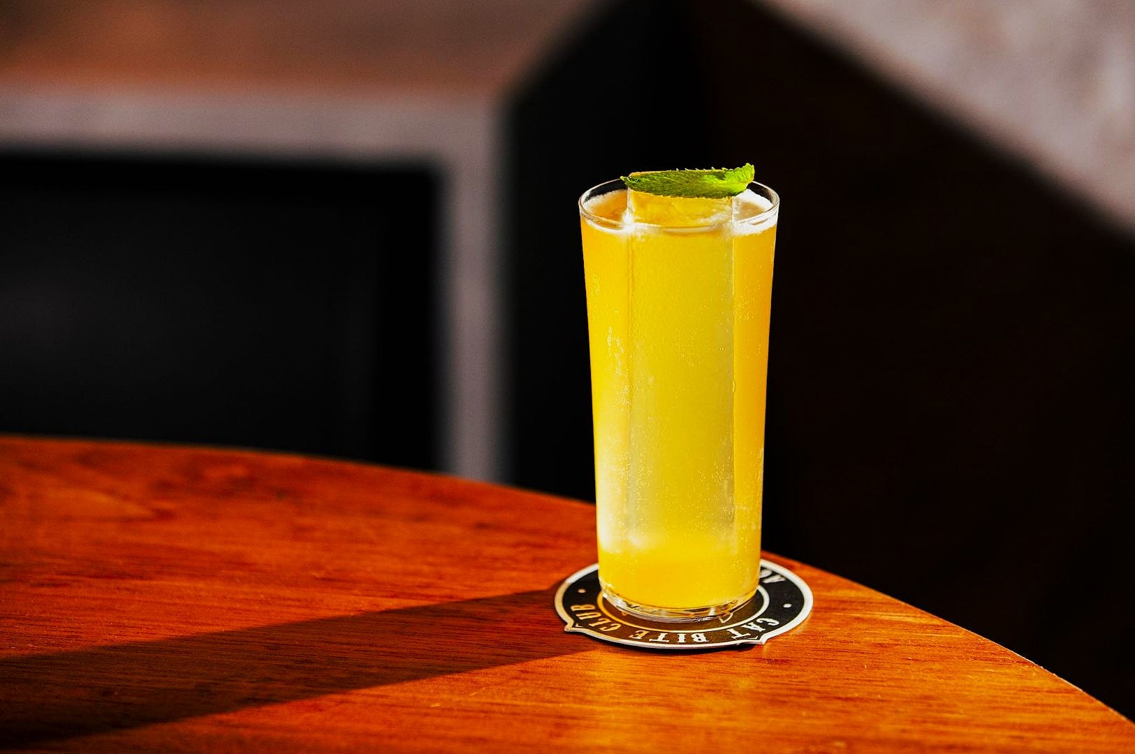 A tall glass of yellow-orange cocktail garnished with a lime wedge and mint leaf sits on a coaster on a wooden table, warmly lit, conveying a refreshing vibe.
