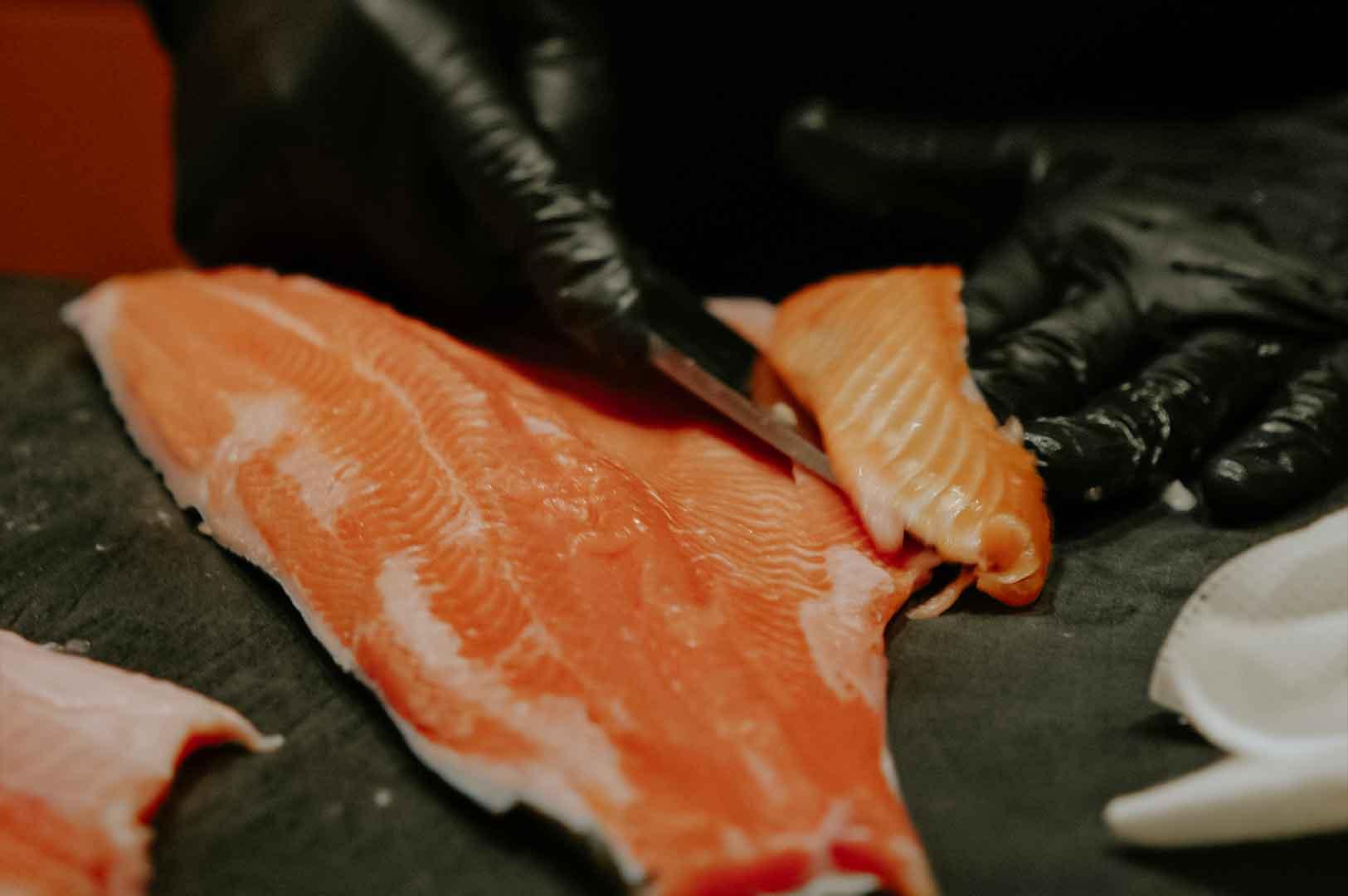 Chef using a sharp knife to cut a fresh salmon fillet into sashimi portions on a dark cutting board.