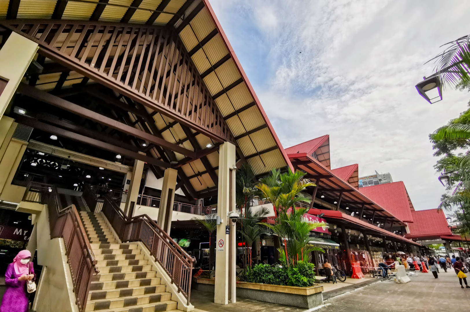 Open-air market with red-roofed buildings, wooden beams, and lush greenery. A person in a pink outfit walks near the stairs under a partly cloudy sky.