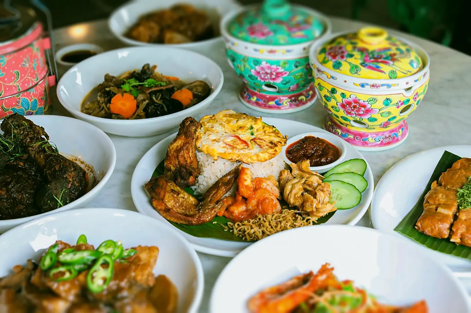 Assortment of Peranakan dishes served on a table, including ayam buah keluak, sambal prawns, braised meats, omelette rice, and colorful traditional tiffin carriers.