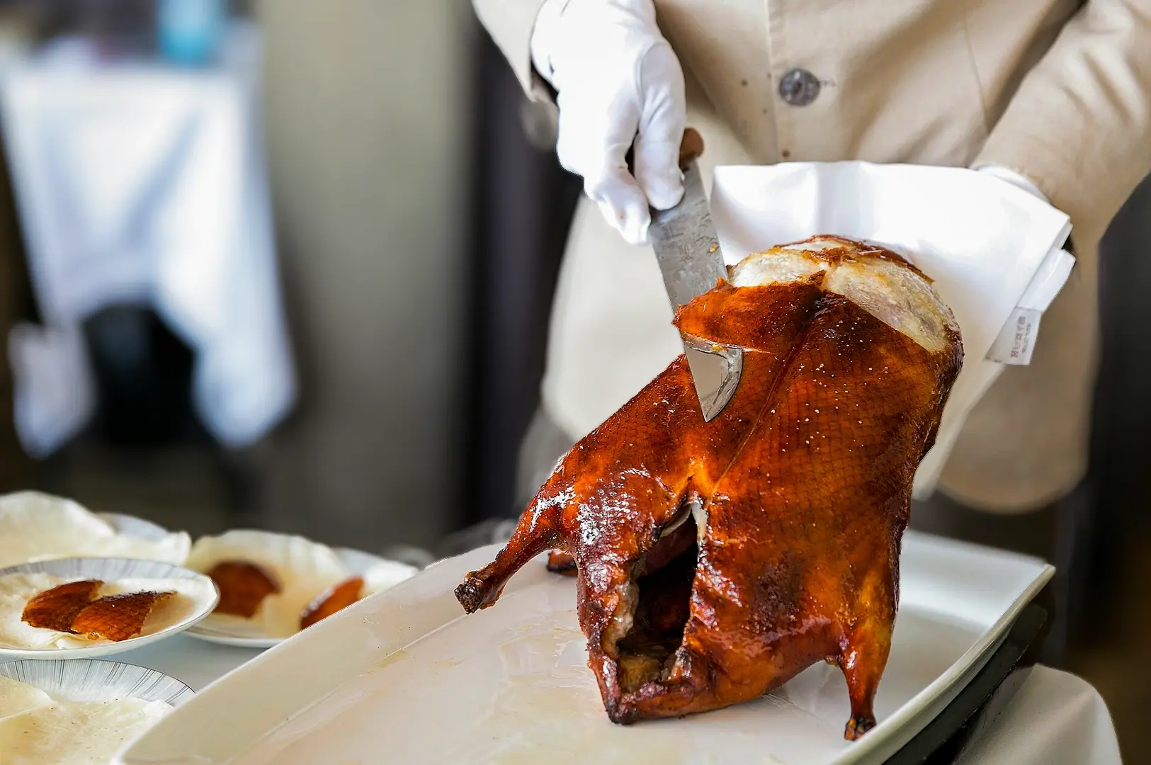 Chef carving a whole roasted Peking duck tableside, slicing the crispy golden skin onto a serving platter with pancakes and dipping sauces.
