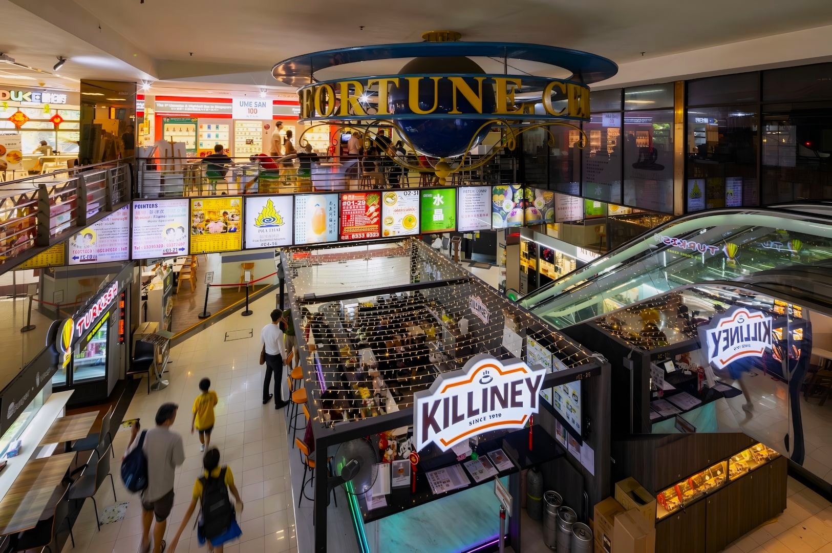 A bustling, multi-level shopping mall interior shows a prominent blue circular Fortune sculpture overhead, bright illuminated menu boards, storefronts, and groups of shoppers navigating the upper and lower levels.