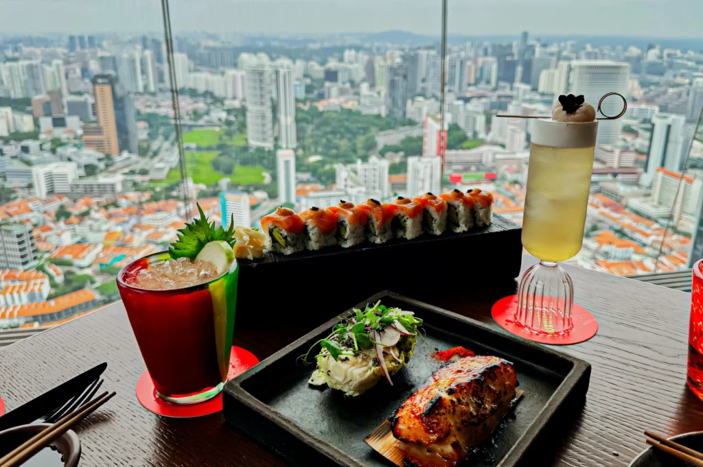 Eye‑level wide shot of a rooftop dining table featuring sushi rolls, grilled entrée, and crafted cocktails in the foreground, with a panoramic city skyline and high‑rise buildings visible through floor‑to‑ceiling windows, highlighting upscale urban dining and skyline views.