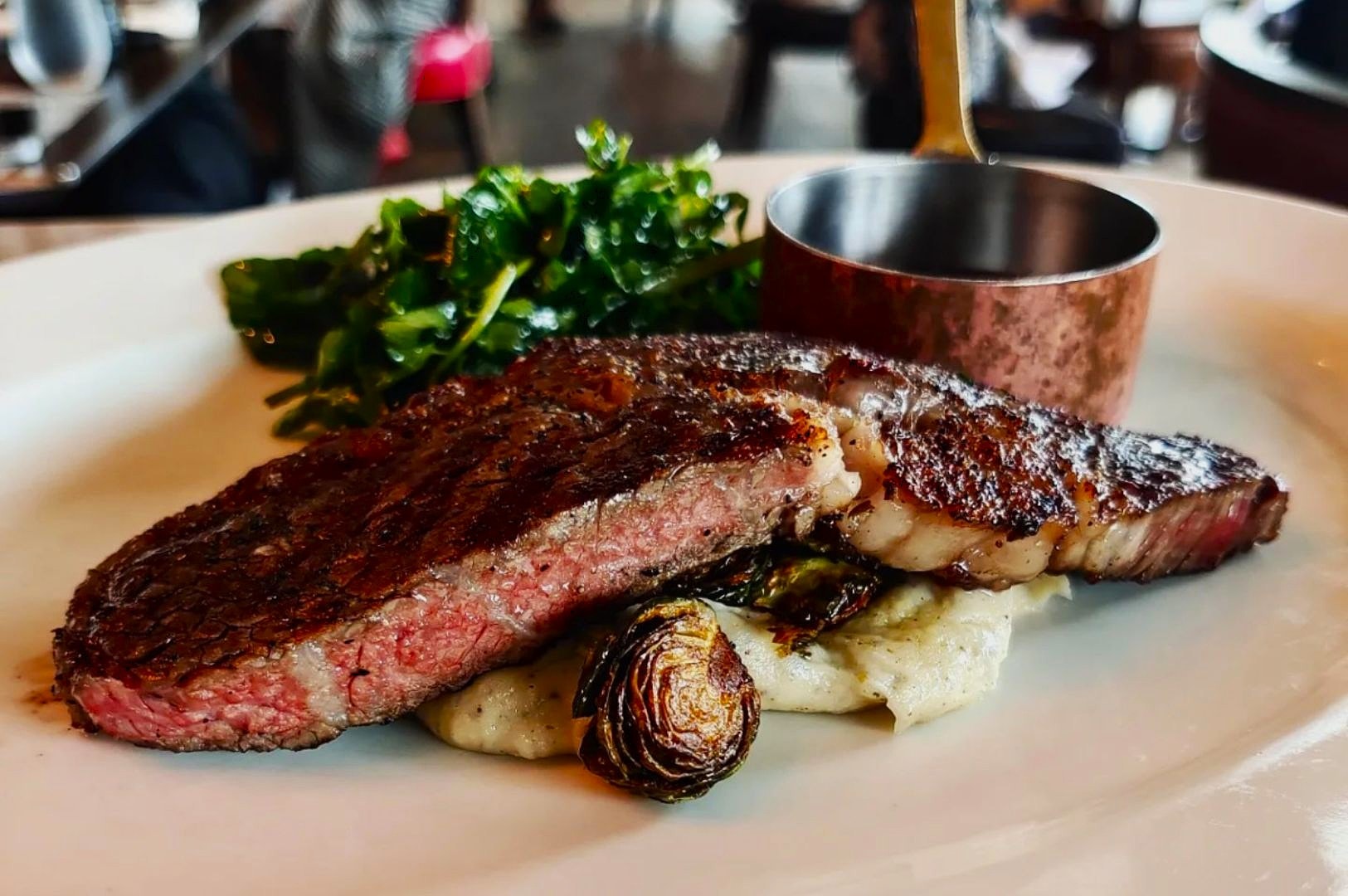 A juicy medium-rare steak sits atop mashed potatoes with charred sprouts, garnished with leafy greens. A small copper saucepot is beside on a white plate.
