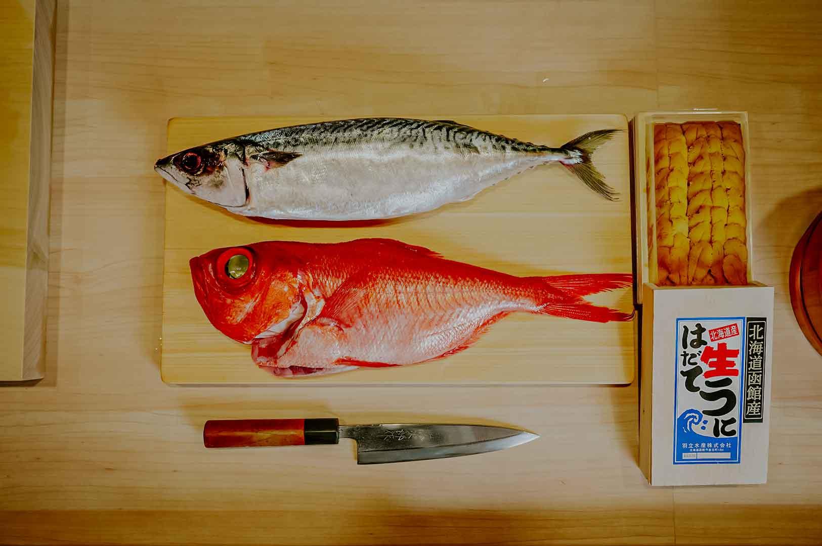 Whole mackerel, red snapper, and a box of fresh uni arranged on a wooden board beside a Japanese chef’s knife at an omakase counter.