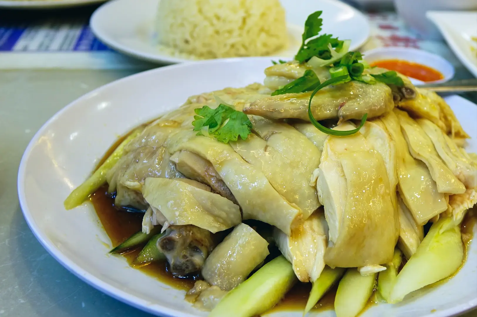 Plate of Hainanese chicken rice with tender poached chicken, sliced cucumbers, soy dressing, and a serving of fragrant rice in the background.
