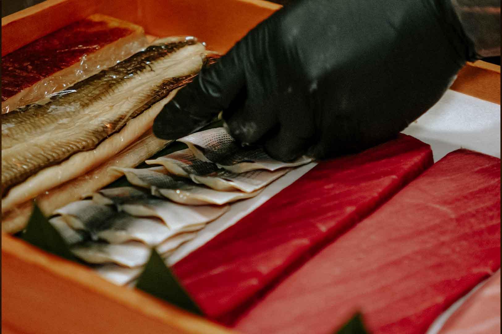 Close-up of assorted premium sashimi cuts including mackerel, tuna, and eel being prepared by a chef wearing black gloves.