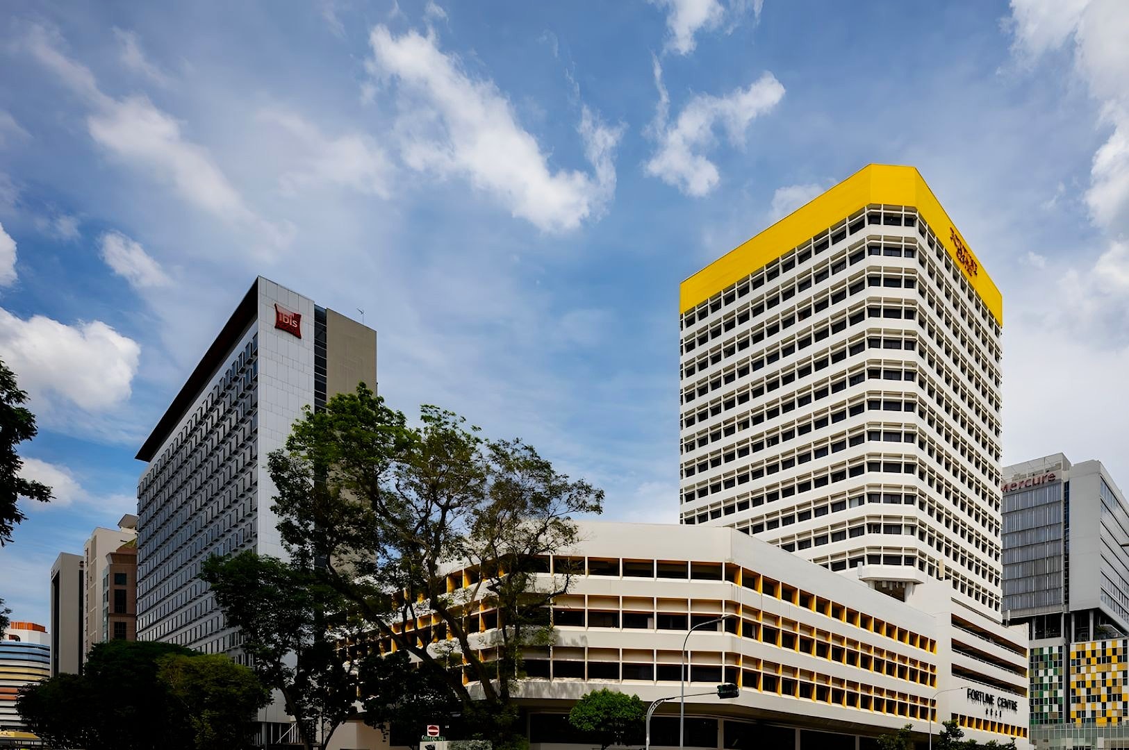 A cityscape shows two dominant mid-rise buildings against a bright blue sky: a white, rectangular hotel-like tower with a red logo and a taller tower topped in bold yellow, surrounded by trees and lower structures.