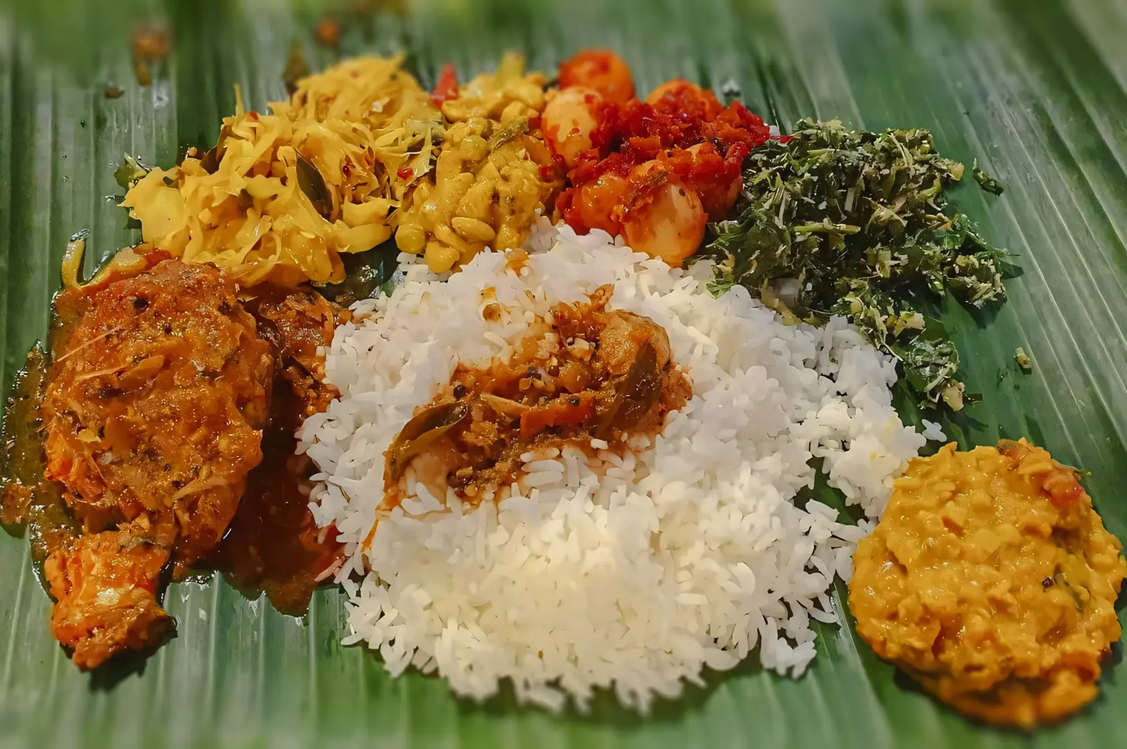 South Indian–style banana leaf meal featuring steamed rice surrounded by spicy chicken curry, assorted vegetables, sambal eggs, greens, and lentil dhal.