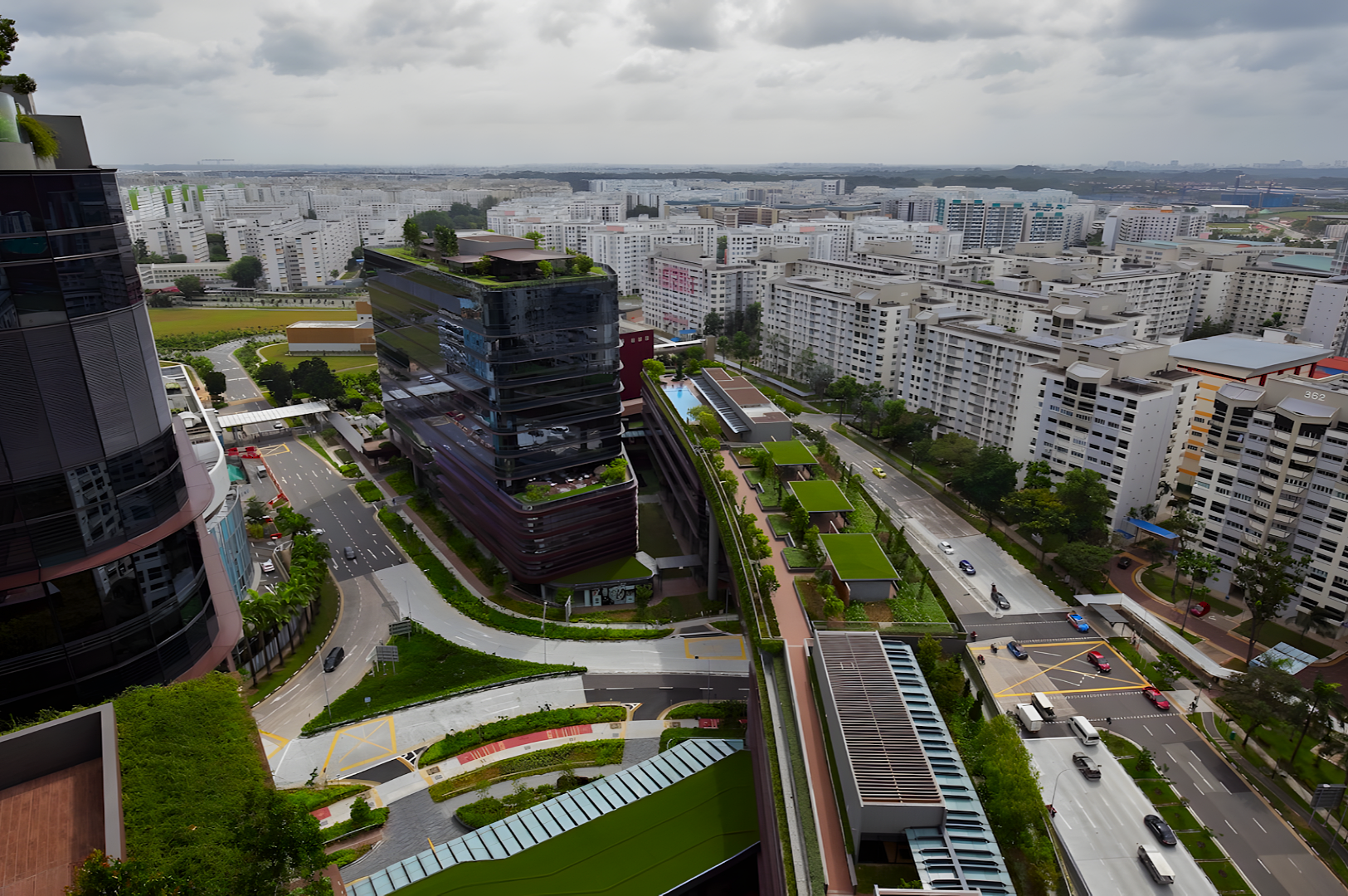 Aerial view of a modern urban landscape featuring tall buildings with green rooftops, surrounding roads, and numerous high-rise apartments under a cloudy sky.