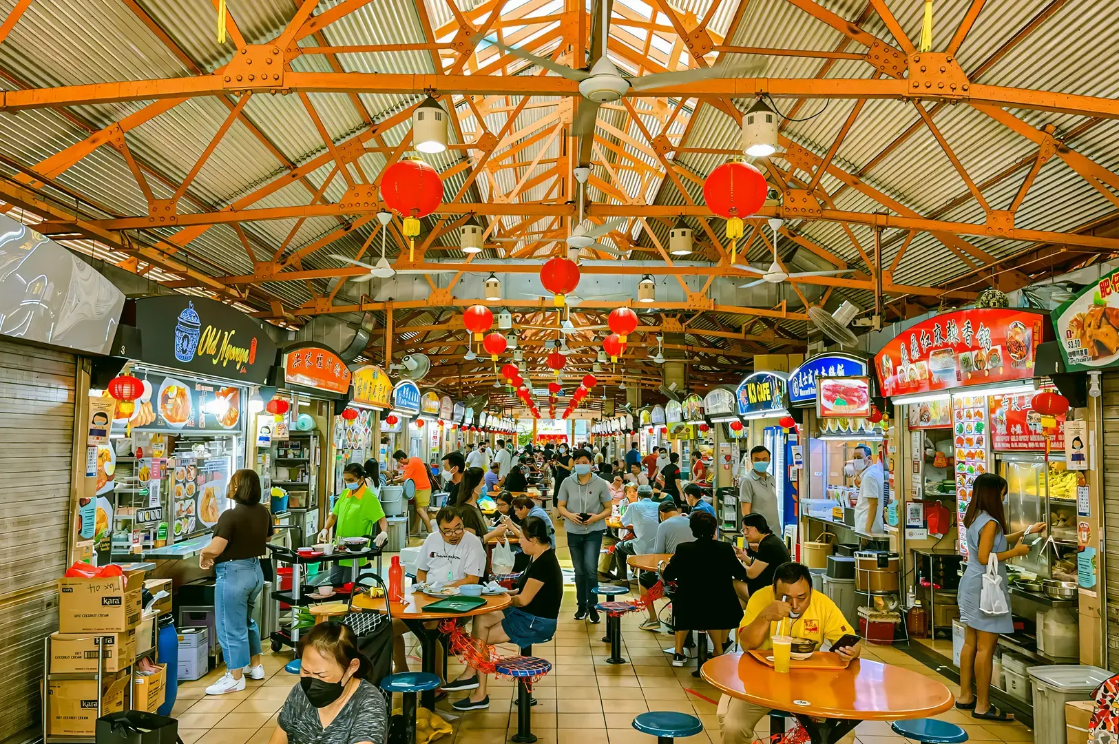 Vibrant Singapore hawker centre featuring food stalls with colorful signboards, festive red lanterns, and diners seated at round tables.