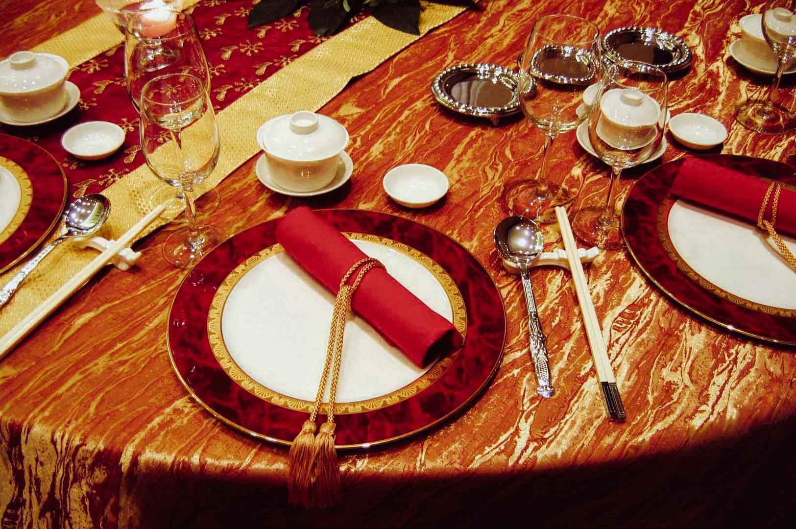 Elegant banquet table setting with red and gold plates, rolled red napkins with tassels, white porcelain dishes, chopsticks, polished silverware, and crystal glassware arranged on a richly textured red and gold tablecloth.