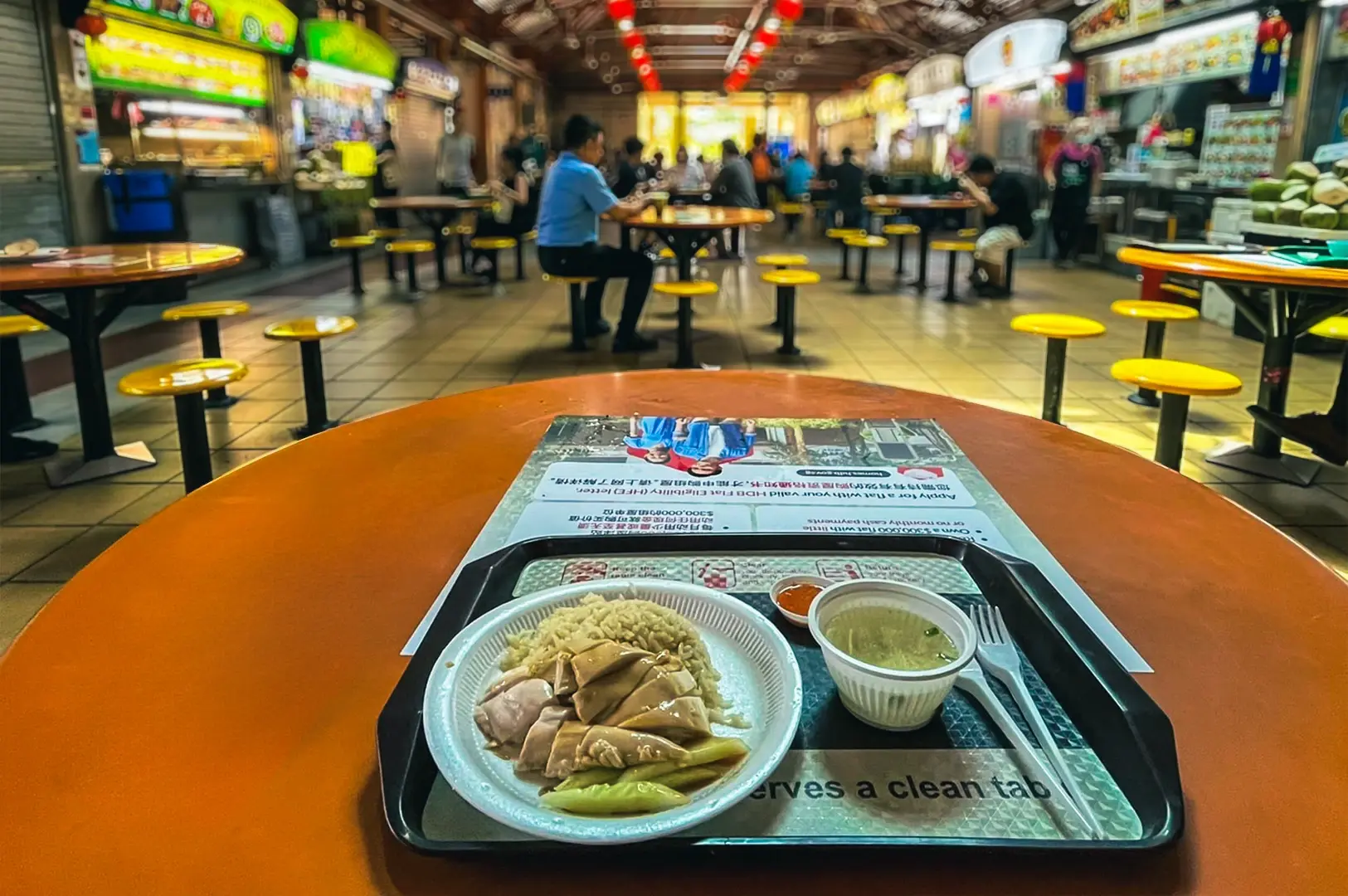 Chicken rice meal on a tray inside a Singapore hawker center with food stalls and tables in the background.