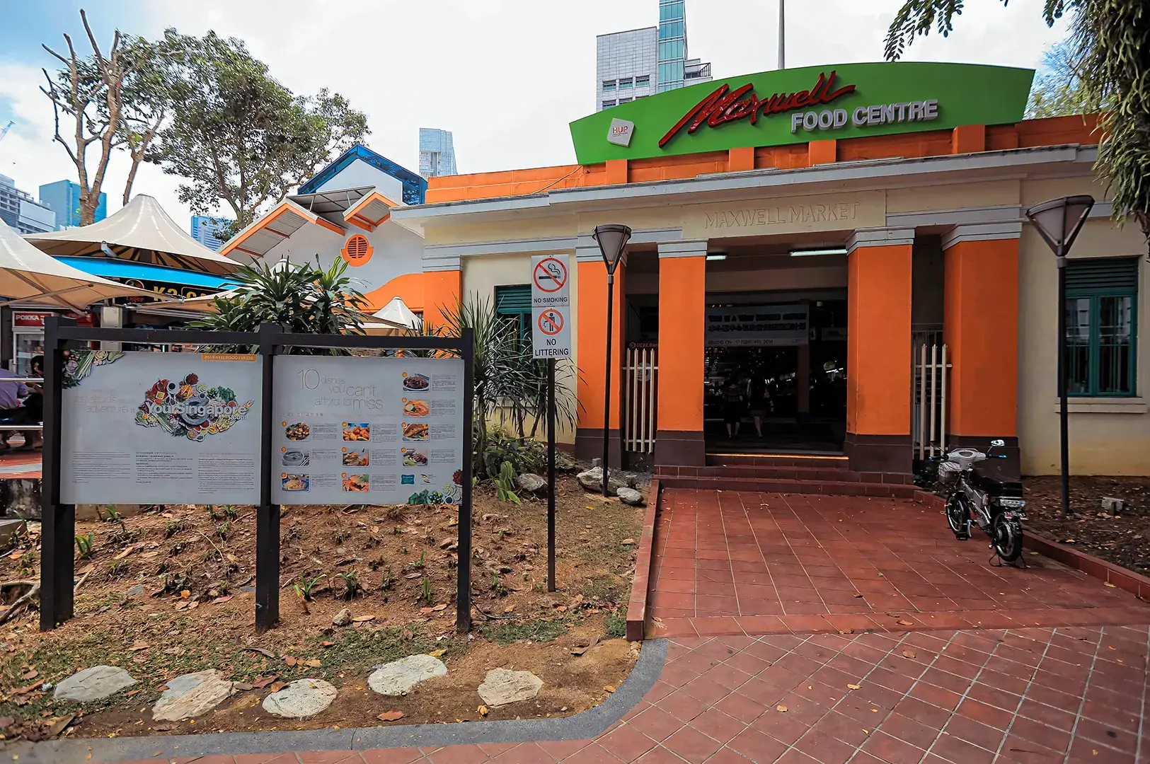 Front entrance of Maxwell Food Centre in Singapore featuring its orange-and-white facade, green signage, outdoor information boards, and surrounding greenery.