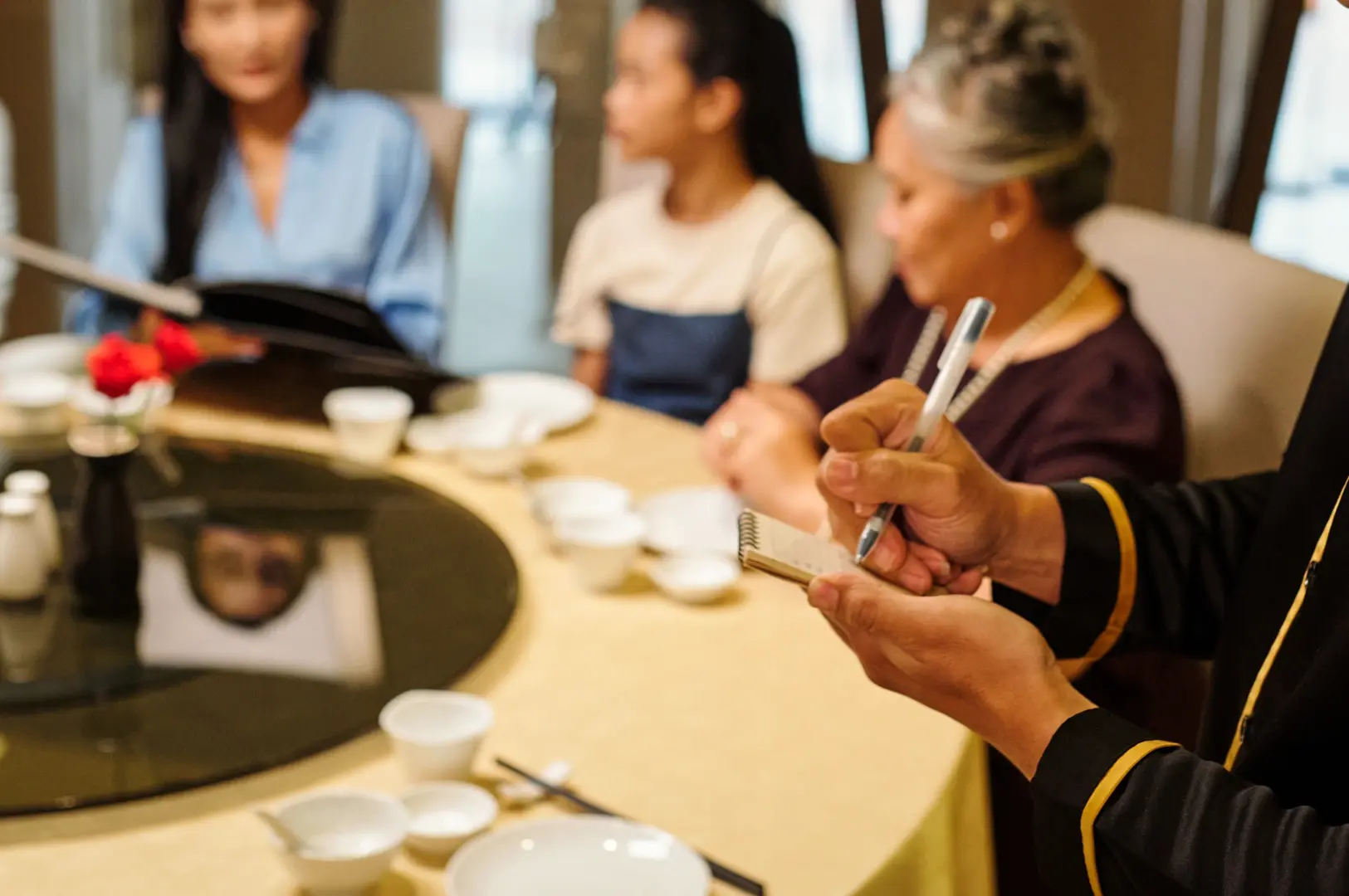 Server writing down a table’s order at a round dining table set with porcelain teacups, and small plates in an elegant Chinese restaurant.