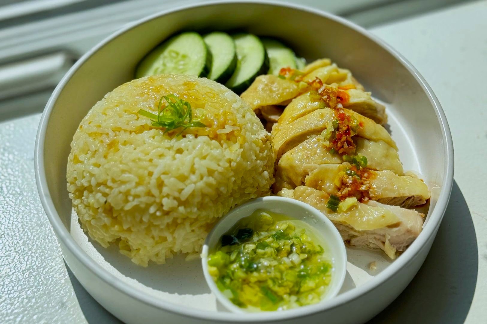 Close view of a plated meal featuring a rice dome, herb-flecked chicken slices with garnished heat, cucumber slices, and a dipping sauce studded with green onions and herbs.