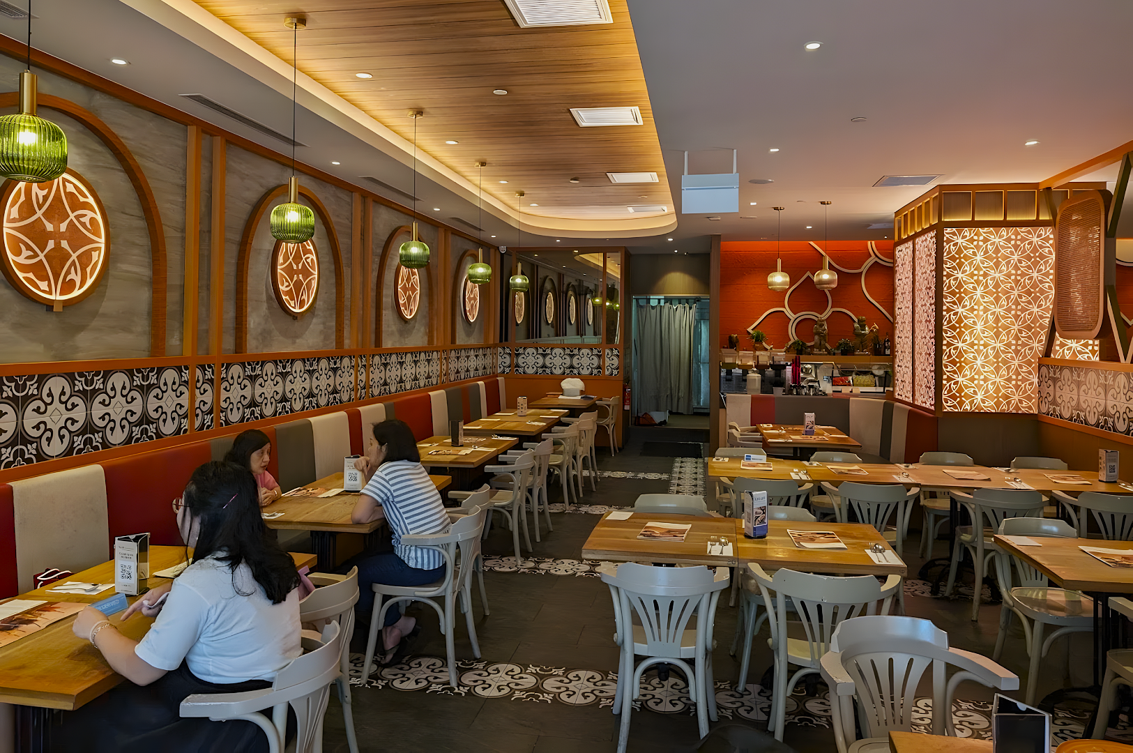 A cozy, modern restaurant interior with wooden tables and white chairs. Warm lighting, ornate wall designs, and pendant lamps create an inviting atmosphere.