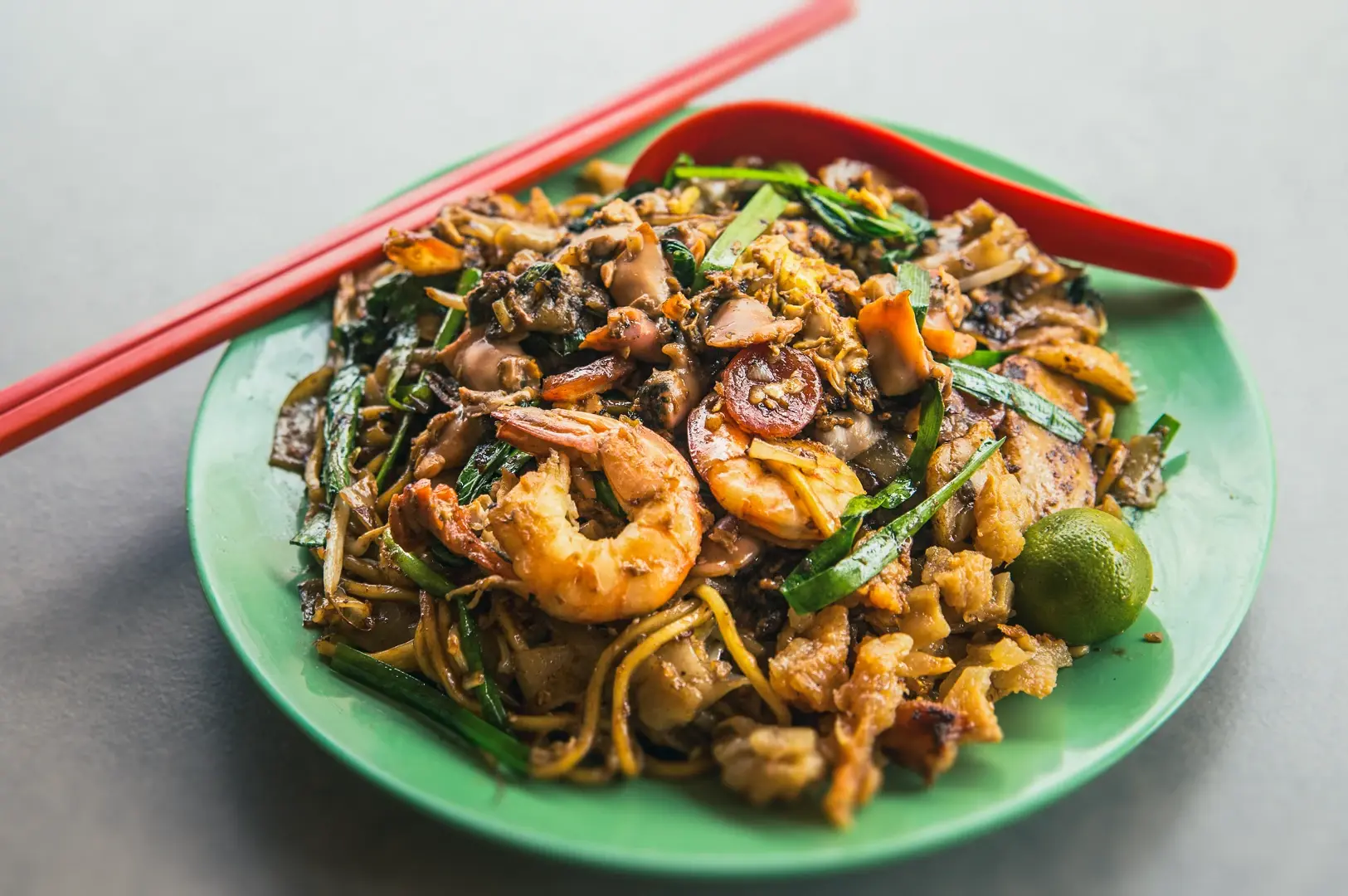 Plate of Singapore Char Kway Teow stir-fried flat rice noodles with prawns, Chinese sausage, egg, bean sprouts, and chives, served with a lime on a green plate.