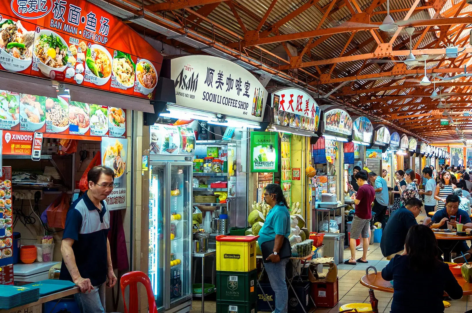 Busy Singapore hawker centre lined with food stalls displaying colorful menu boards, refrigerators, cooking stations, and customers dining at plastic tables.