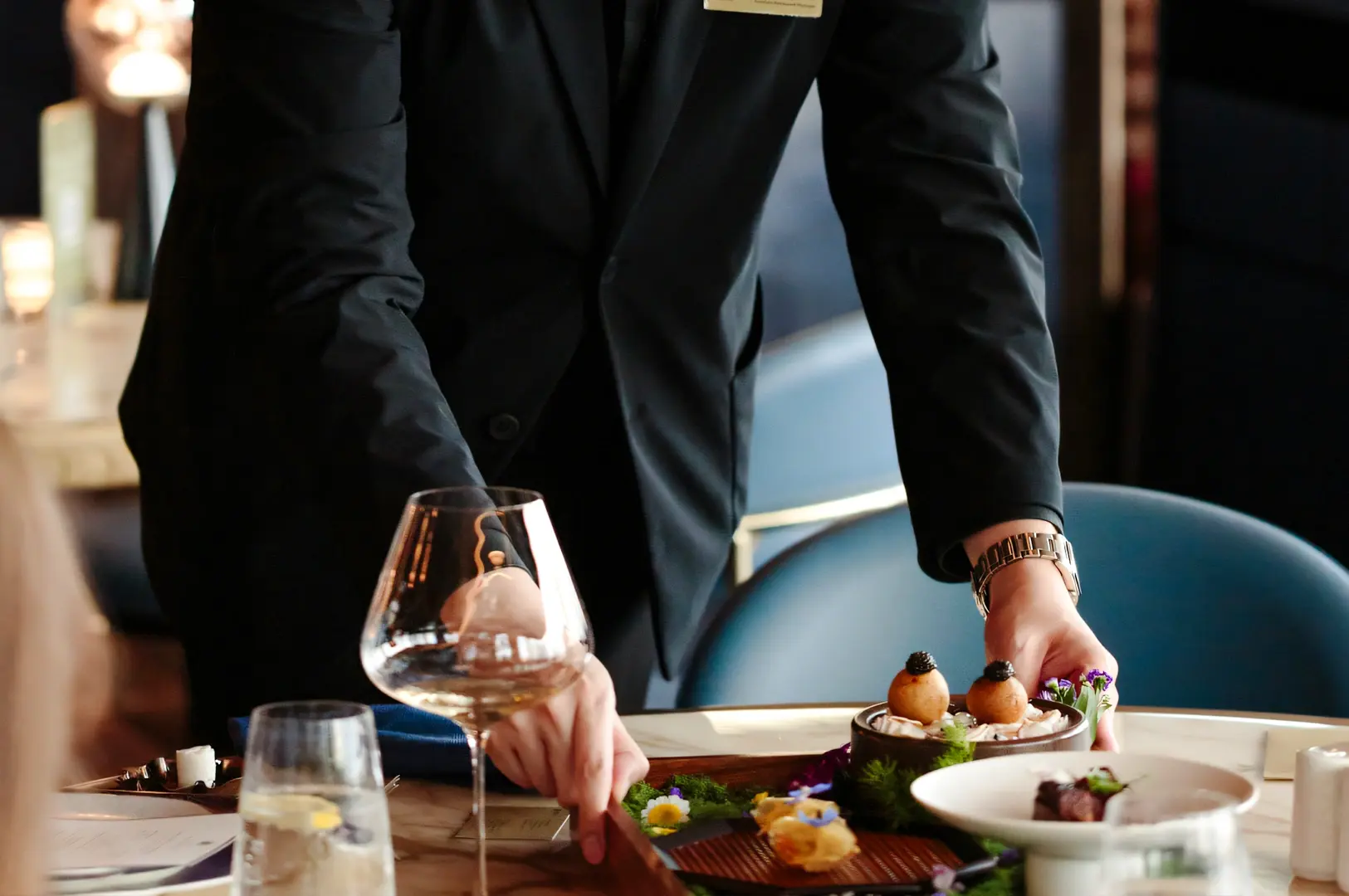 Server in formal attire presenting an elegant fine‑dining dish with caviar‑topped appetizers and artistic plating on a restaurant table set with wine glasses and modern décor.