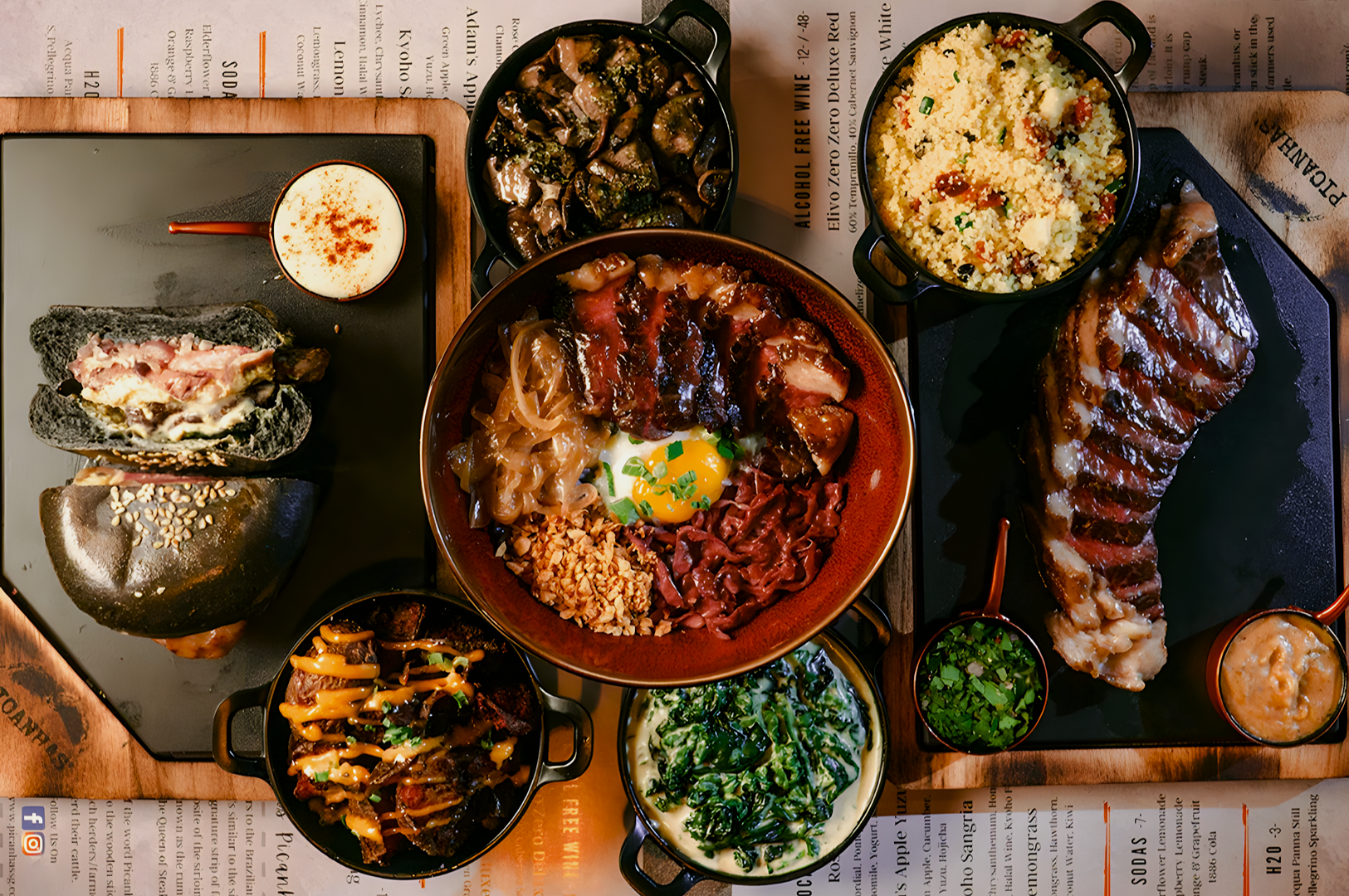 An assortment of gourmet dishes on a table including a charcoal bun burger, grilled meat bowl, steak, and various sides like spinach and couscous.