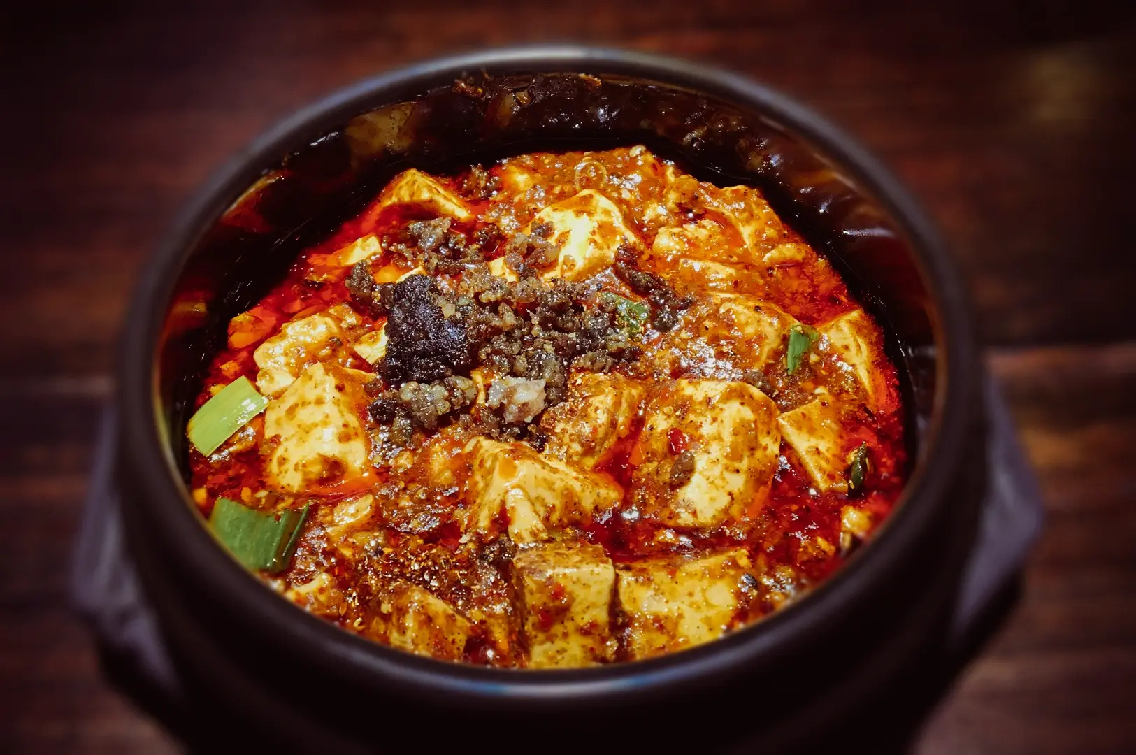 Close‑up of mapo tofu served in a black clay pot, showcasing soft tofu cubes in a rich, spicy Sichuan chili sauce topped with minced ingredients.