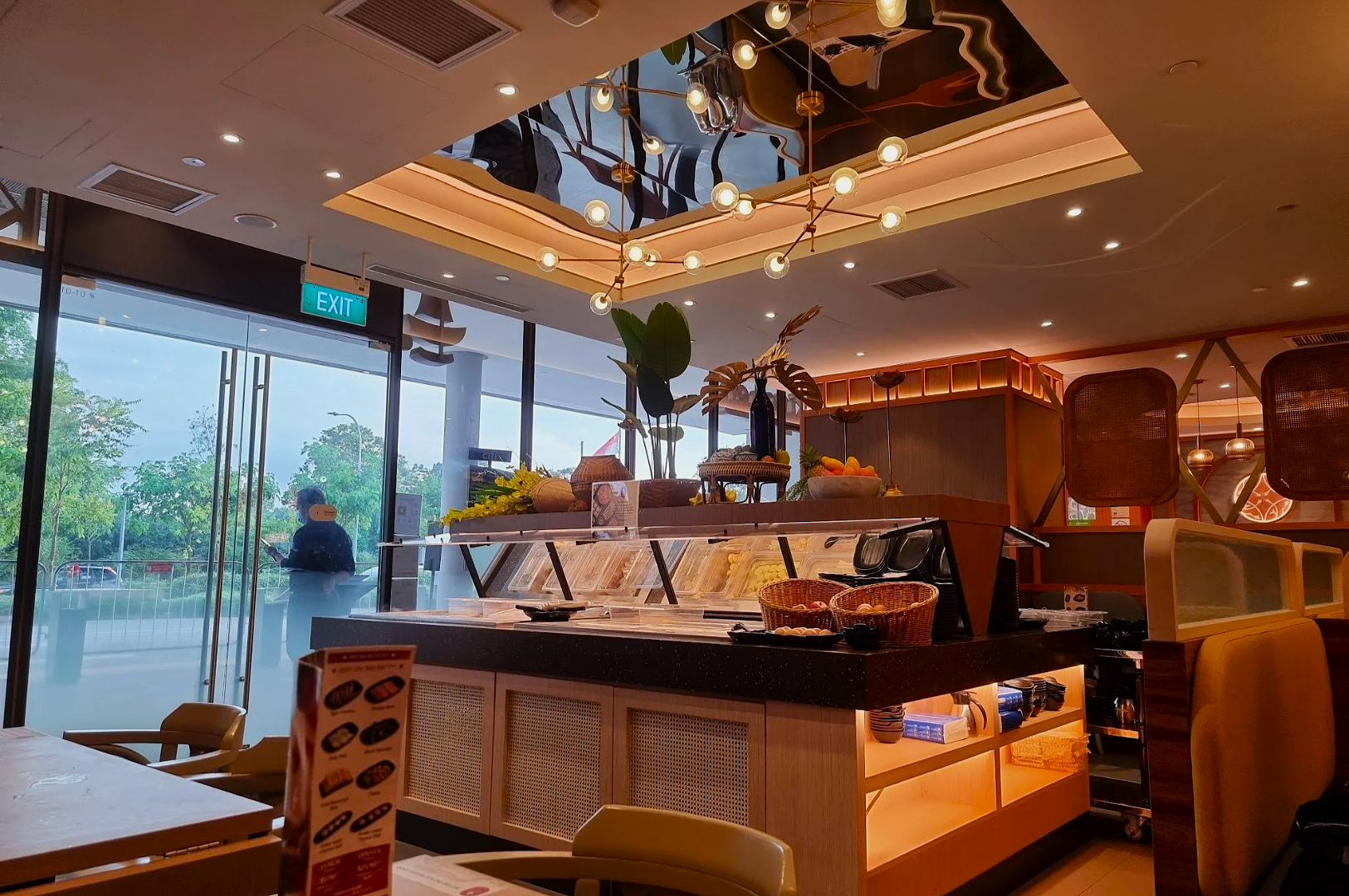 Modern restaurant interior with a stylish ceiling light fixture and decorative plants. Buffet counter with fresh ingredients is in view. Soft, inviting lighting and outdoor greenery create a relaxed atmosphere.