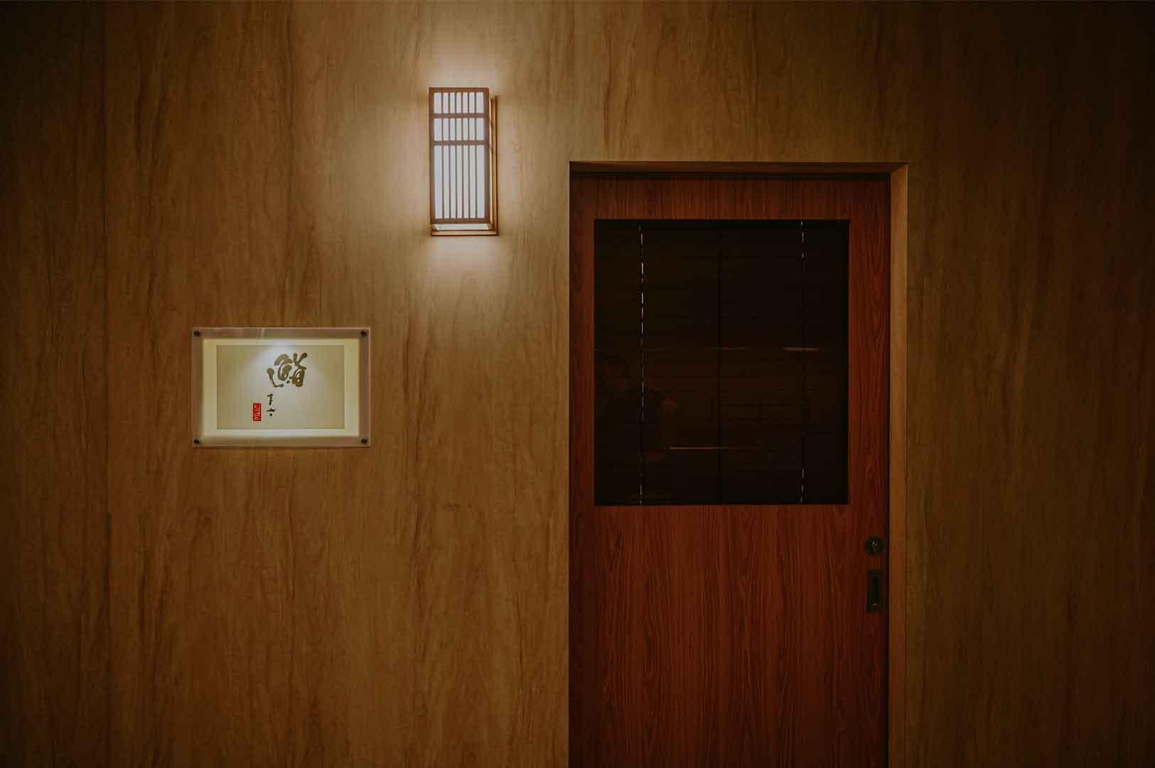 Minimalist Japanese restaurant entrance featuring a wooden door, warm wall lighting, and illuminated signage.