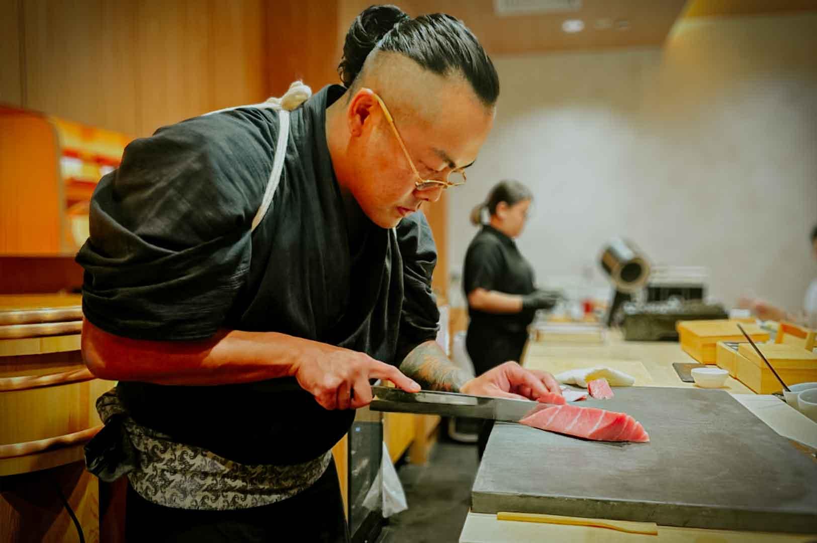 Chef expertly slicing a block of fresh tuna on a stone board inside a traditional Japanese omakase kitchen.